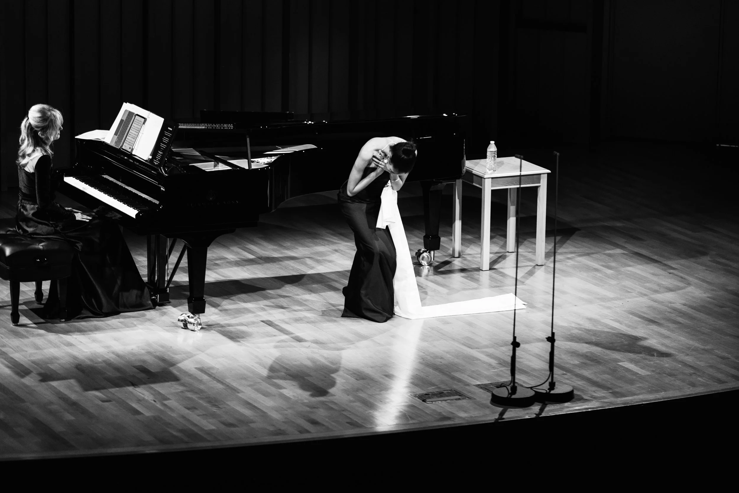 A stage with a female pianist and a woman in an emotional pose near a grand piano, a water bottle on a small table, and microphones in the foreground, captured in black and white.