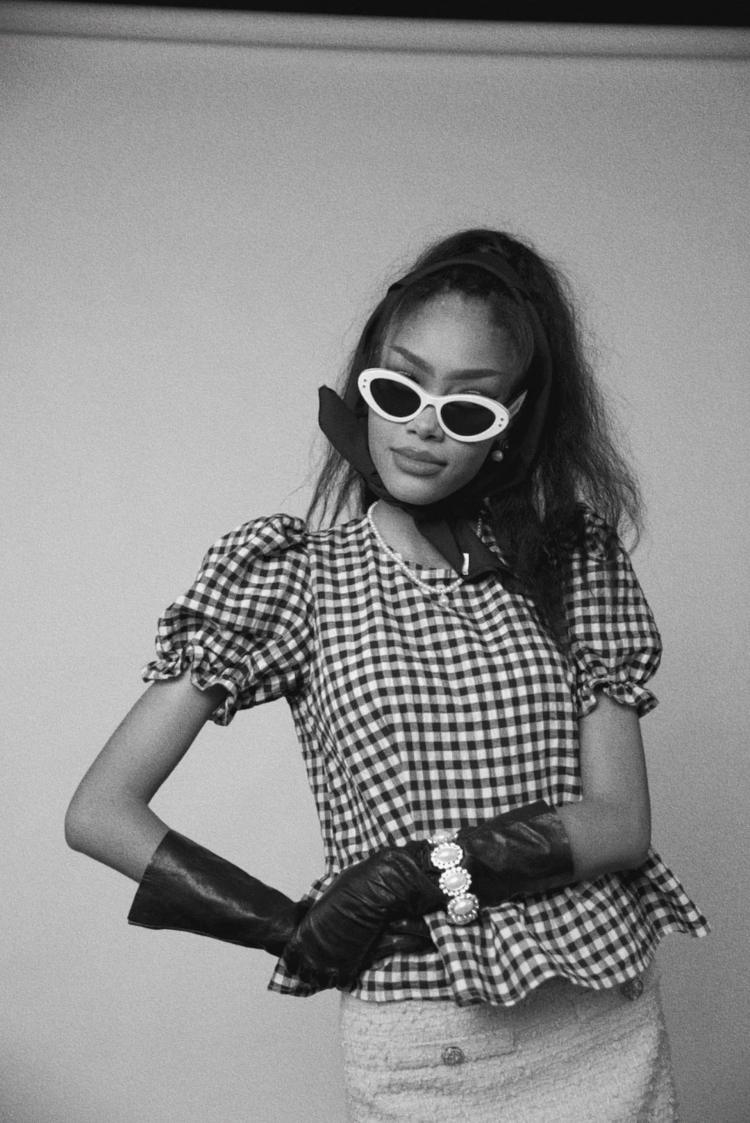 Opera Singer and voice actor Randall Gray with dark hair styled in a ponytail, wearing a black headband, dressed in a white top with black accents, layered pearl jewelry, and makeup with bold eyeliner, posing against a plain background.