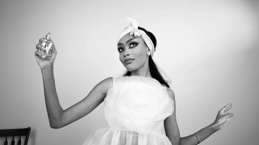Black and white photo of opera singer and voice actor, Randall Gray, wearing a headscarf, strapless top, and floral earrings, with makeup and hair styled in vintage fashion, against a plain background.