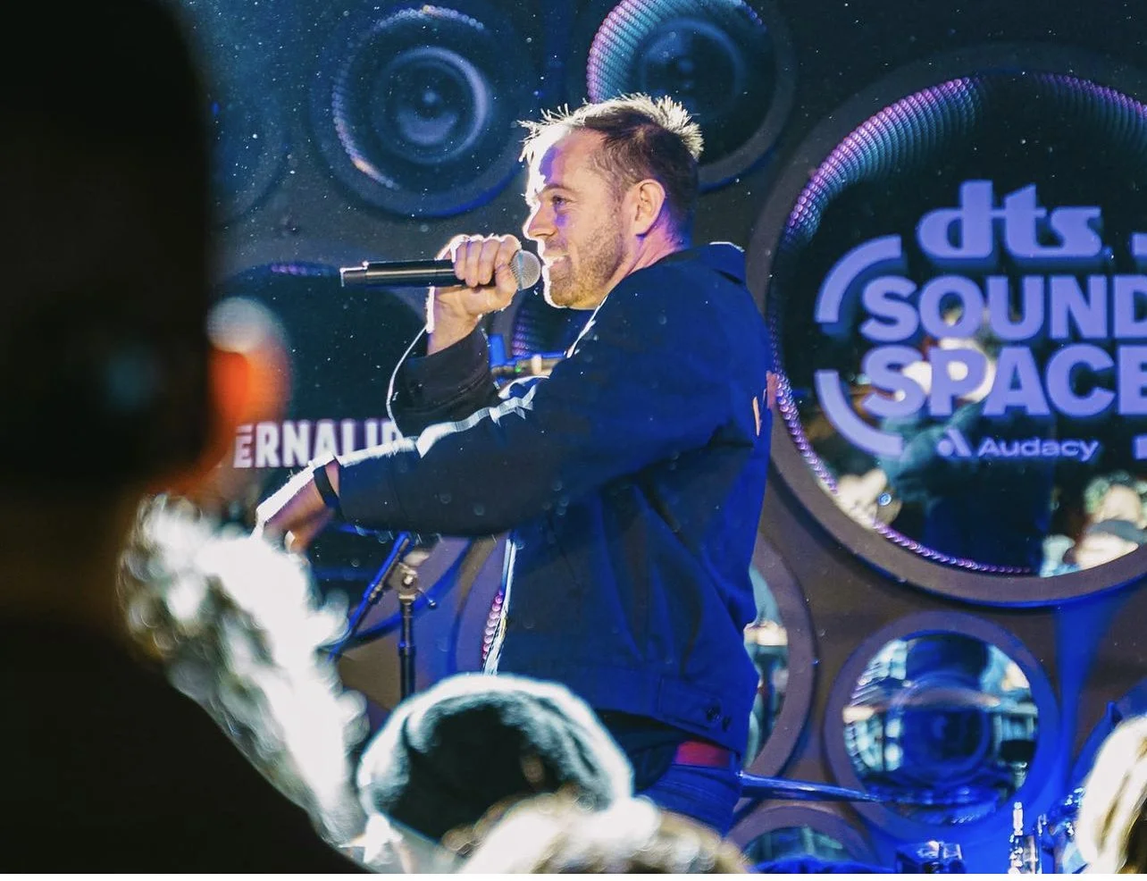 A man holding a microphone performing on stage at a sound space event, with a large screen displaying the words "dts SOUND SPACE" in the background.