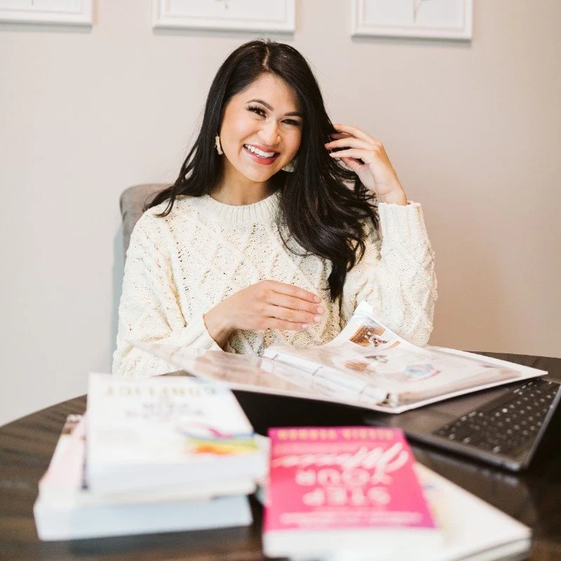A smiling woman with long dark hair, wearing a white sweater, sitting at a desk with magazines, a laptop, and a pink book in front of her.