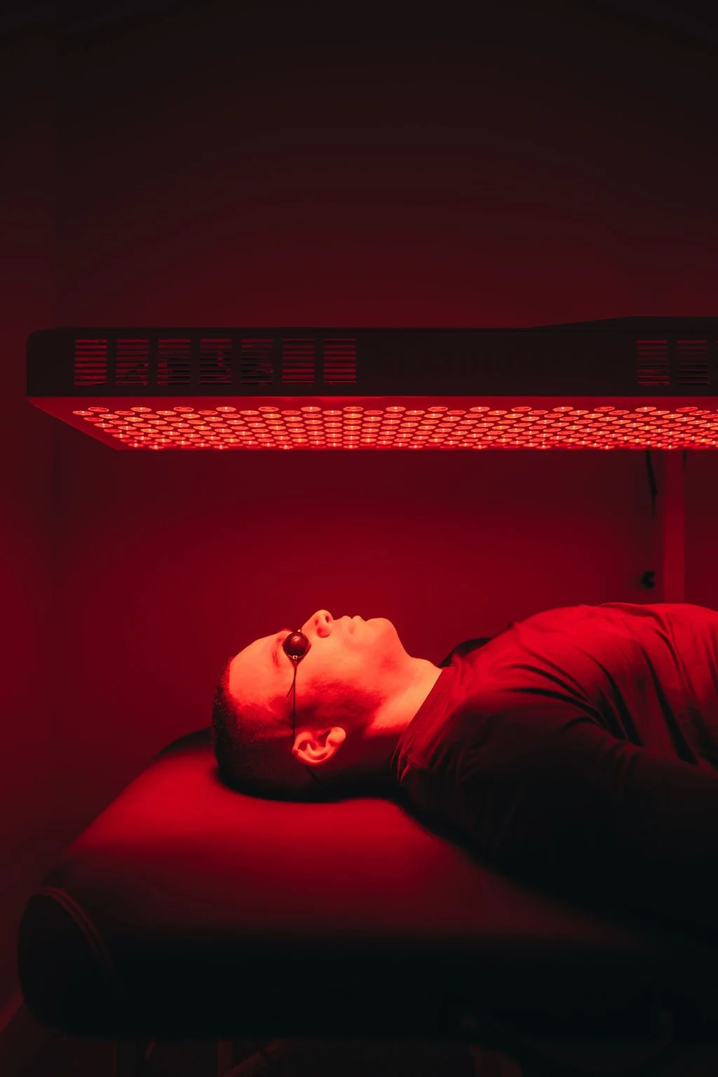 A man lying on a table under a red infrared therapy lamp, wearing protective glasses, in a dimly lit room.