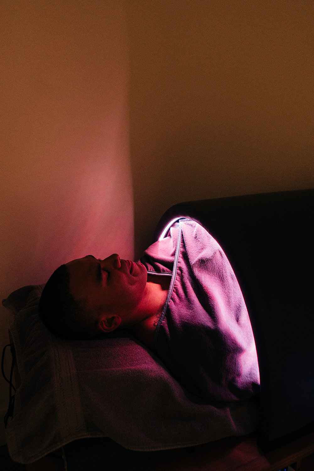 Person lying in a bed with a blanket, illuminated by a pinkish glow from a small, portable LED light.