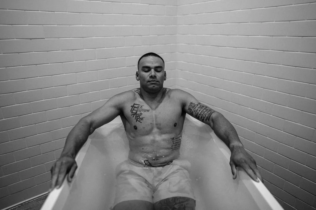 A shirtless man with tattoos sitting in a bathtub filled with water, eyes closed, with brick wall tiles in the background.