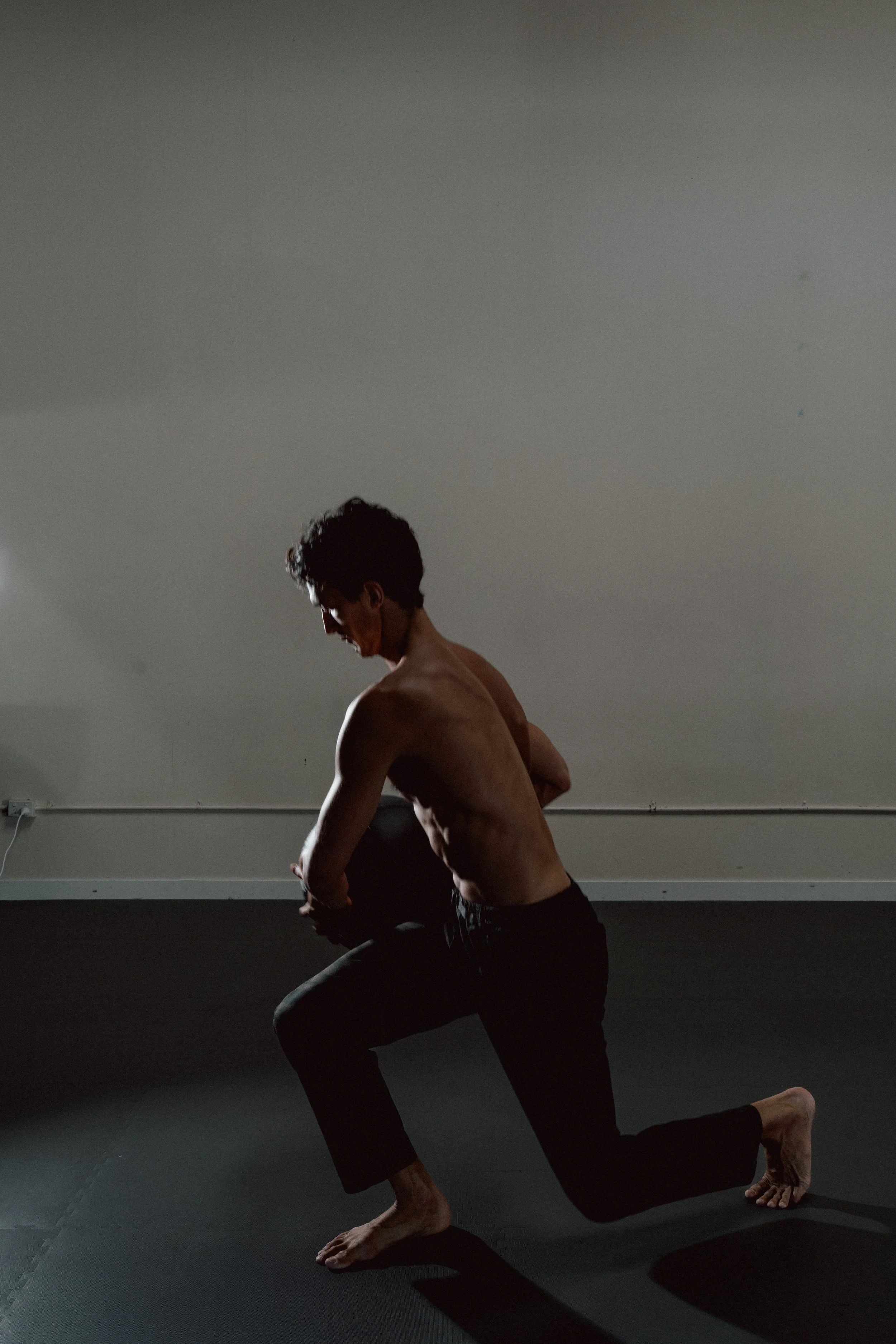 A man practicing yoga on a black mat, balancing on one knee and one hand, with his other arm and leg extended. He's shirtless, wearing dark pants, and is indoors against a plain wall.