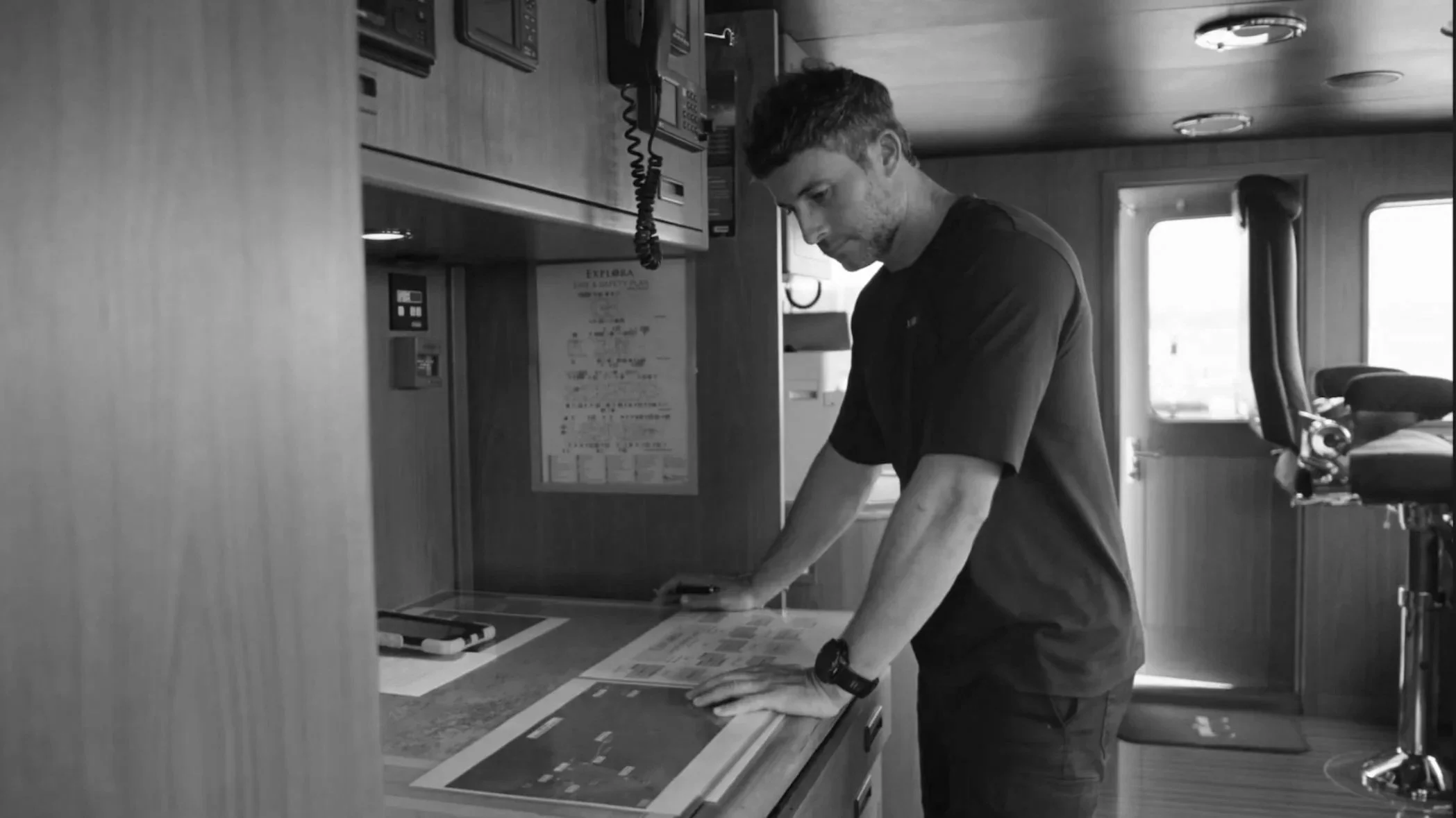 A man with short hair leaning over a counter in a room, possibly an office or control room, with various equipment, a fire safety poster, and a telephone mounted on the wall.