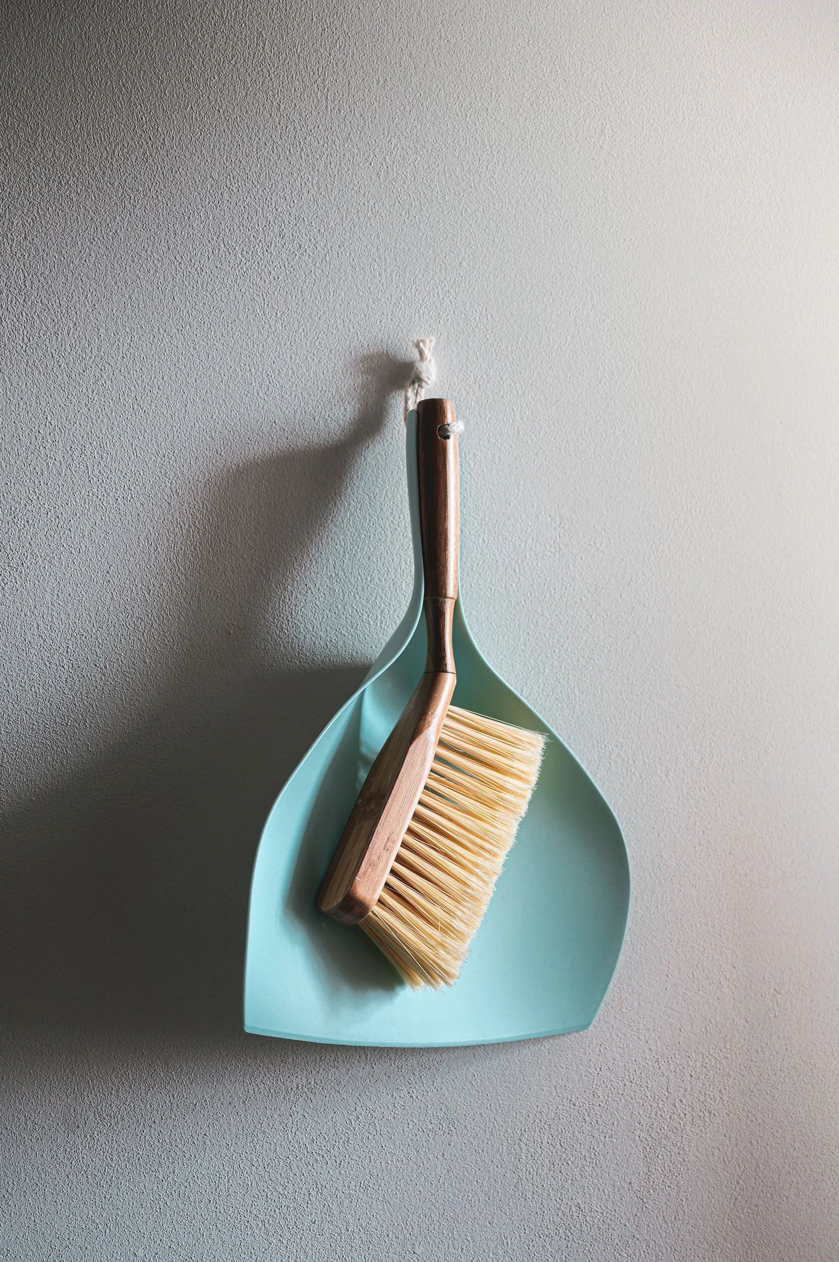 A light blue dustpan with a wooden-handled brush resting inside, hanging on a hook on a textured gray wall.