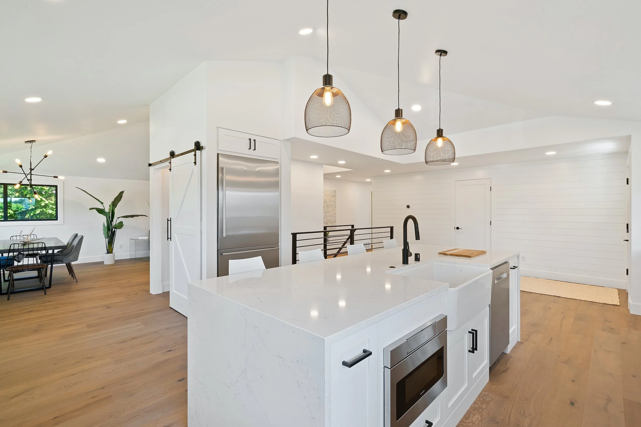 Modern kitchen with white island, black fixtures, and pendant lights, open to dining area with seating and a window with plants outside.