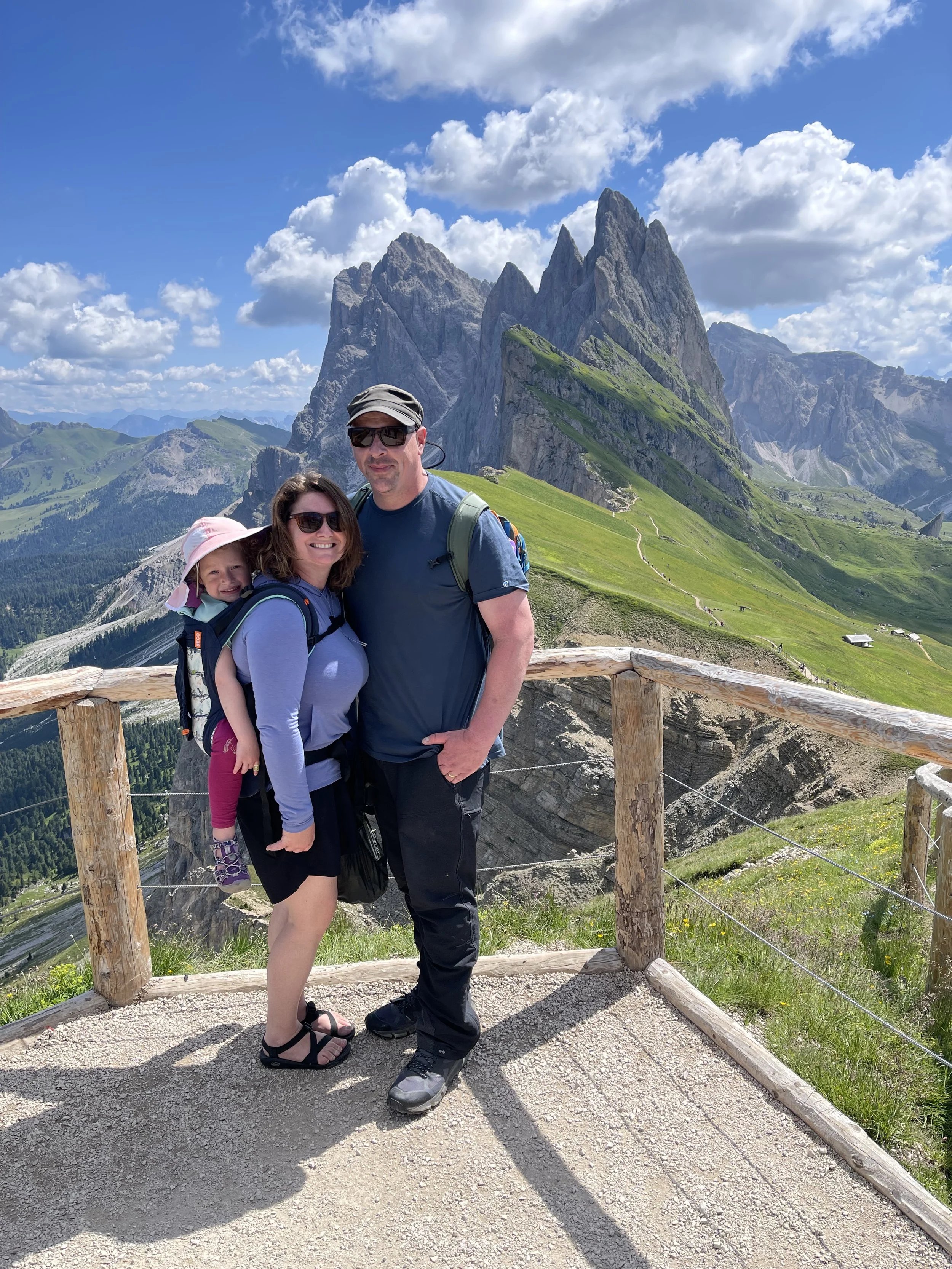 A family of three standing on a wooden viewing platform with mountains and green hills in the background under a partly cloudy sky.