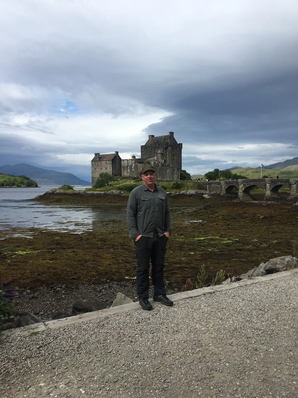A man stands in front of Eilean Donan Castle in Scotland. there is water and clear skies in the background.