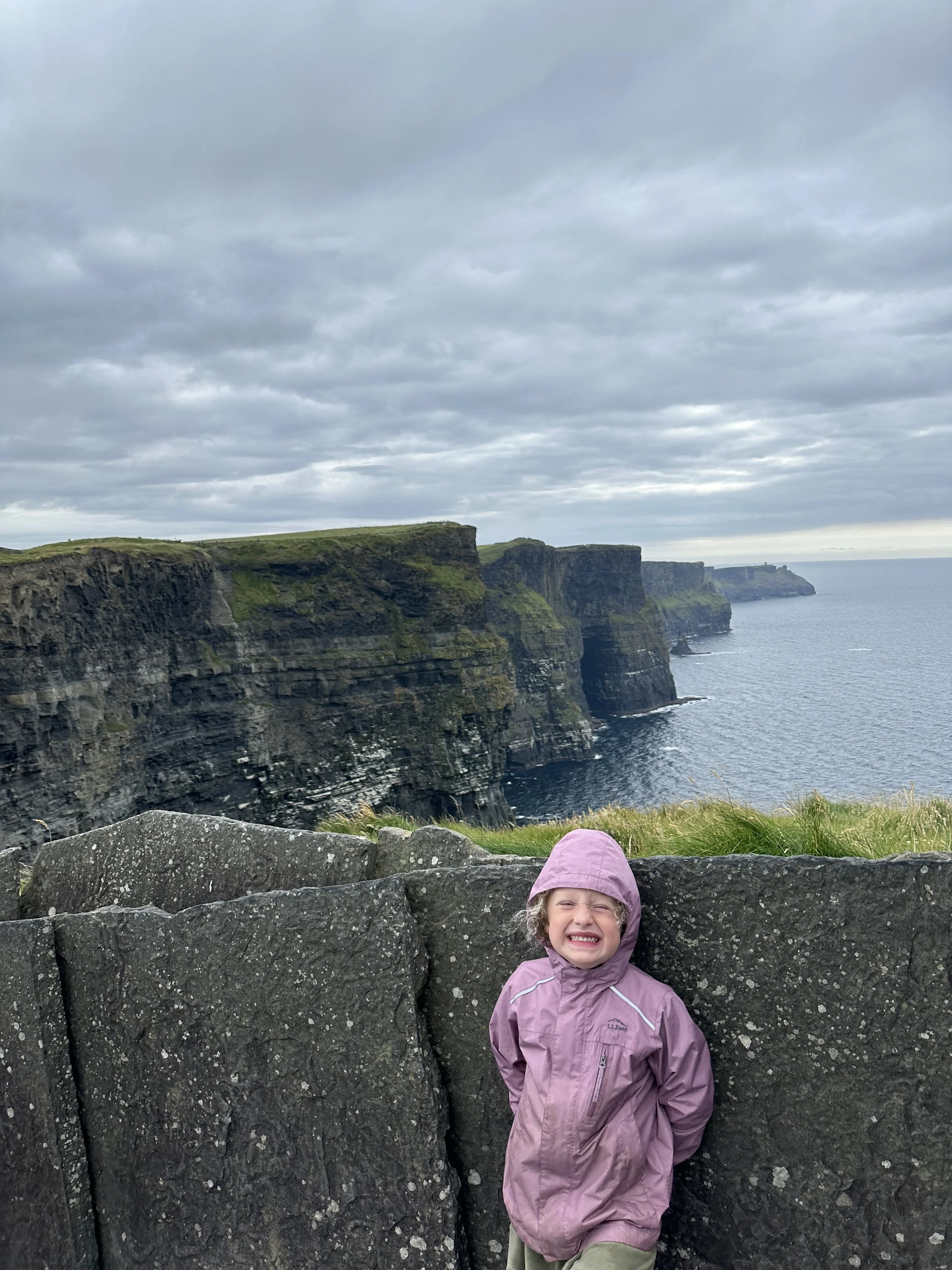 A CHILD STANDS BY THE WALL IN FRONT OF THE CLIFFS OF MOHER. THE ATLANTIC OCEAN IS IN THE BACKGROUND.