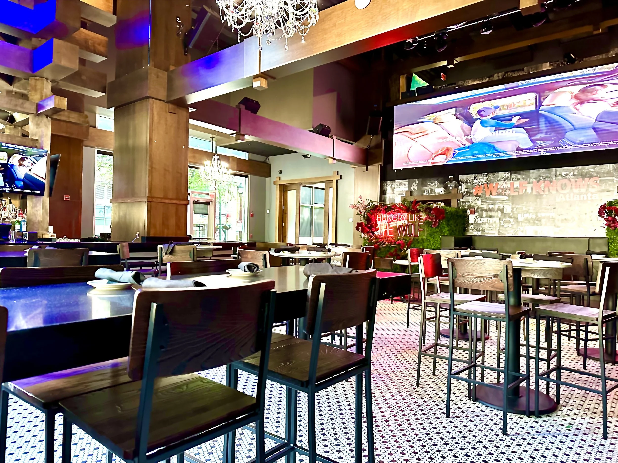 Empty restaurant dining area with wooden tables and chairs, large windows allowing natural light, a chandelier hanging from the ceiling, and multiple large screens displaying content.
