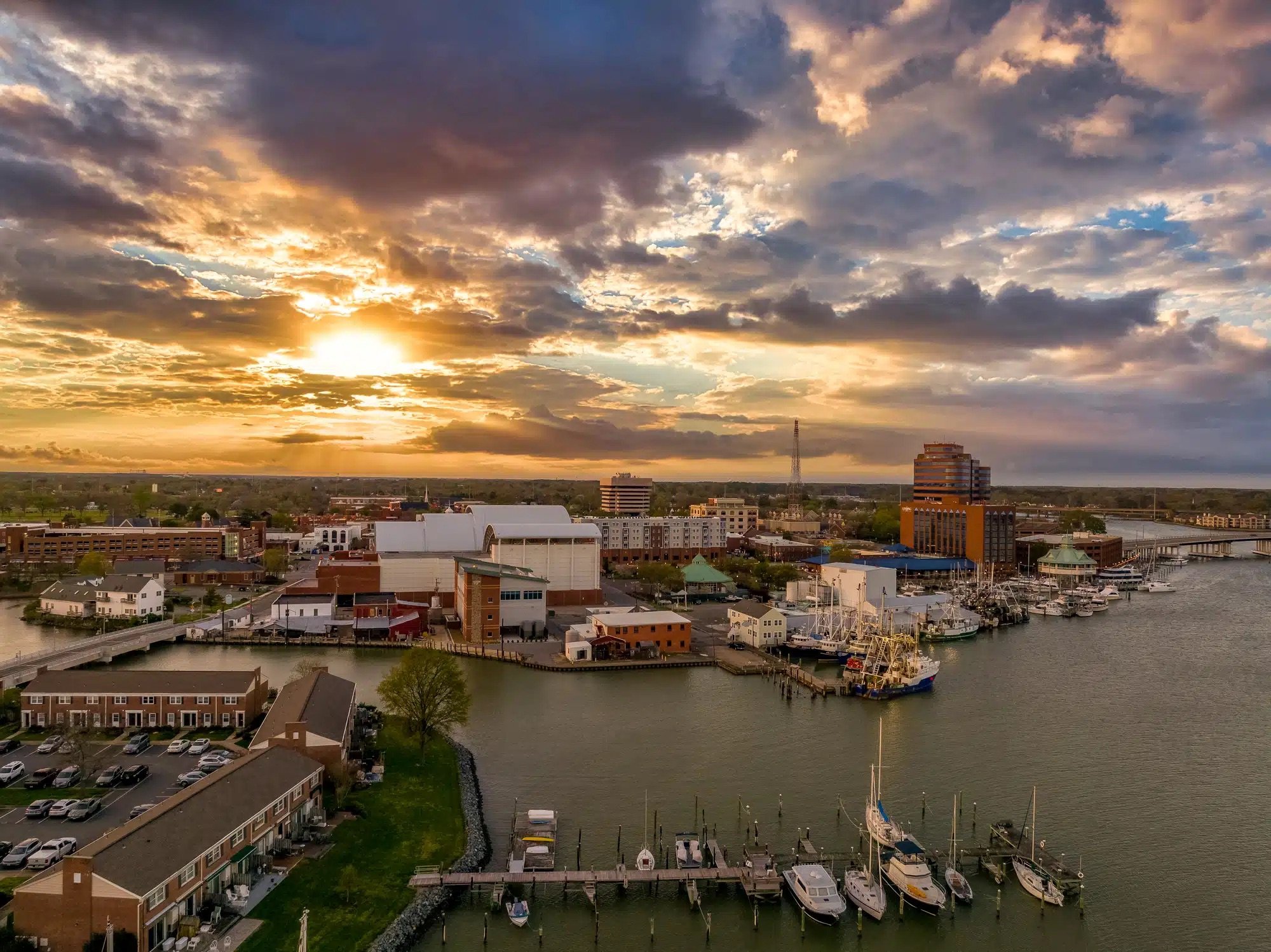 Aerial view of a harbor city at sunset with boats docked, buildings, and dramatic clouds.
