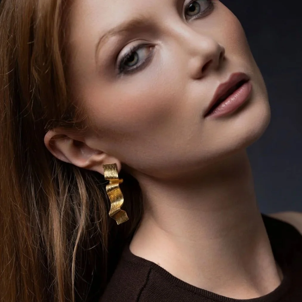 Close-up of a woman with red hair, gold earring, and makeup looking slightly to the side against a dark background.