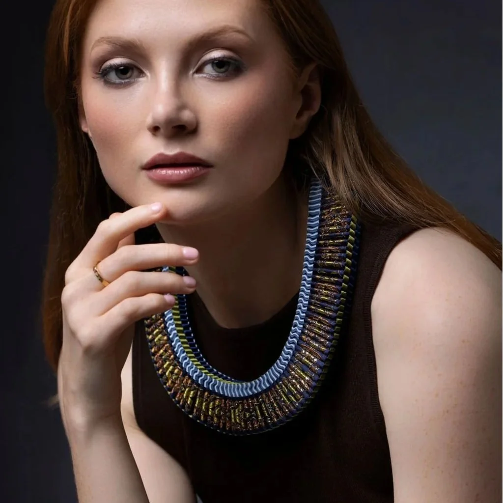 A woman with red hair and fair skin wearing a black sleeveless top and a statement ribbon braided necklace, with her hand resting near her chin, against a dark background.
