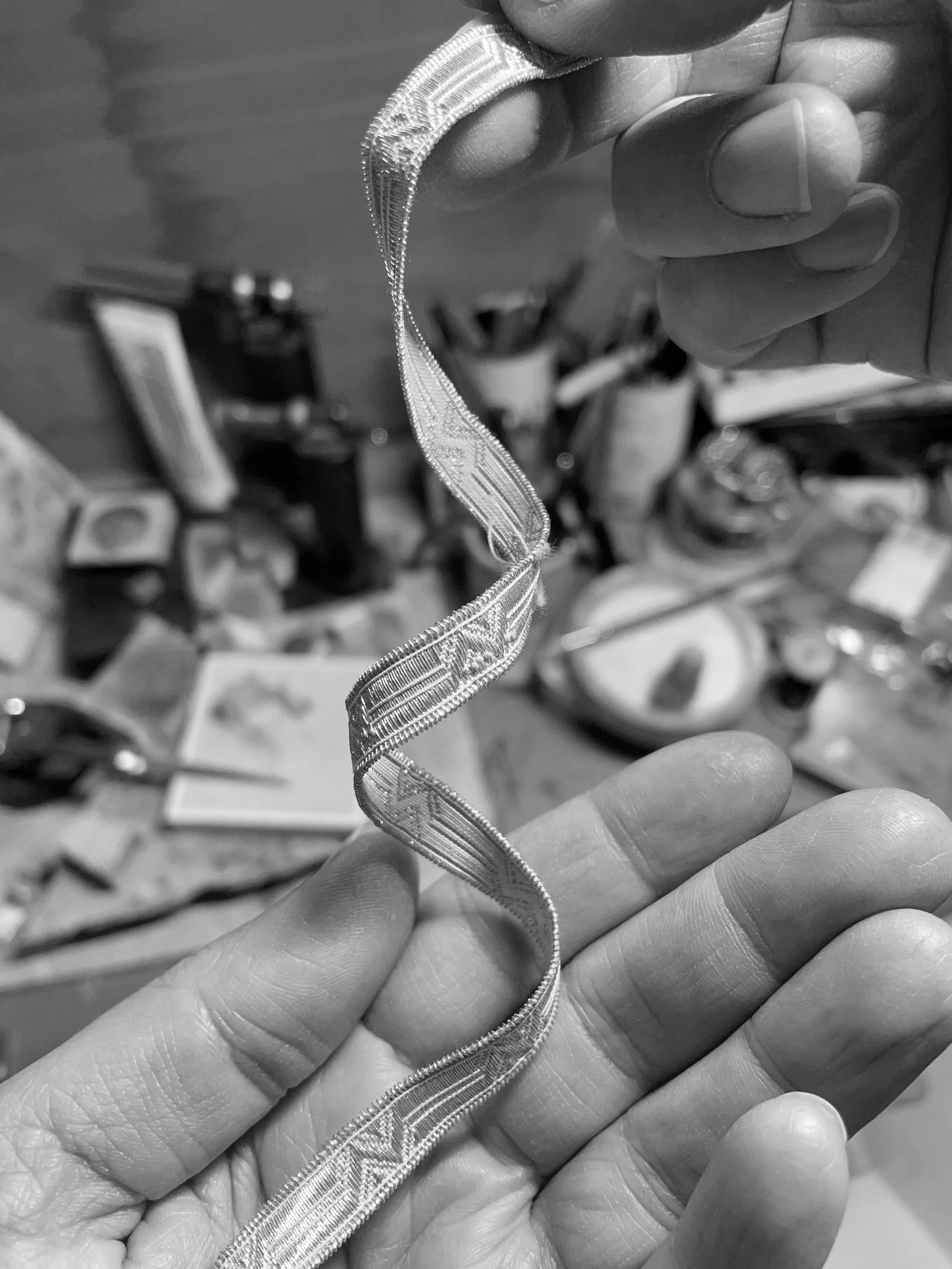 Close-up black and white photo of a person's fingers holding a delicate, patterned silver bracelet, with a blurred workshop background.