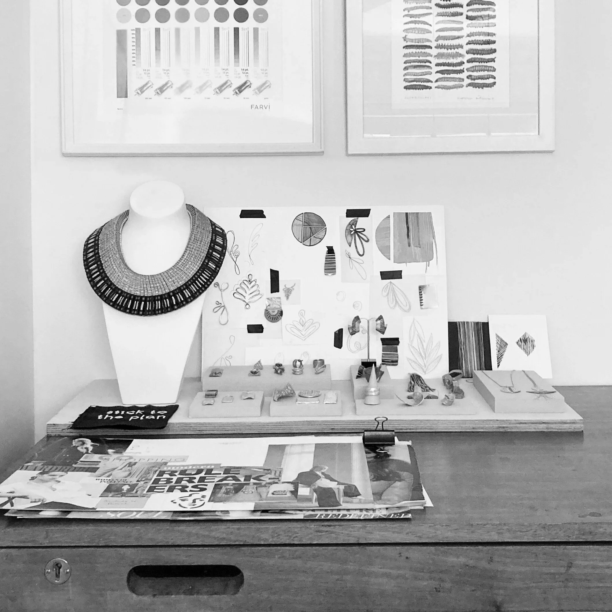 Display of various posters and printed materials on a wall, including a geometric pattern, text and illustrations, with a table below holding stacks of printed magazines or brochures.
