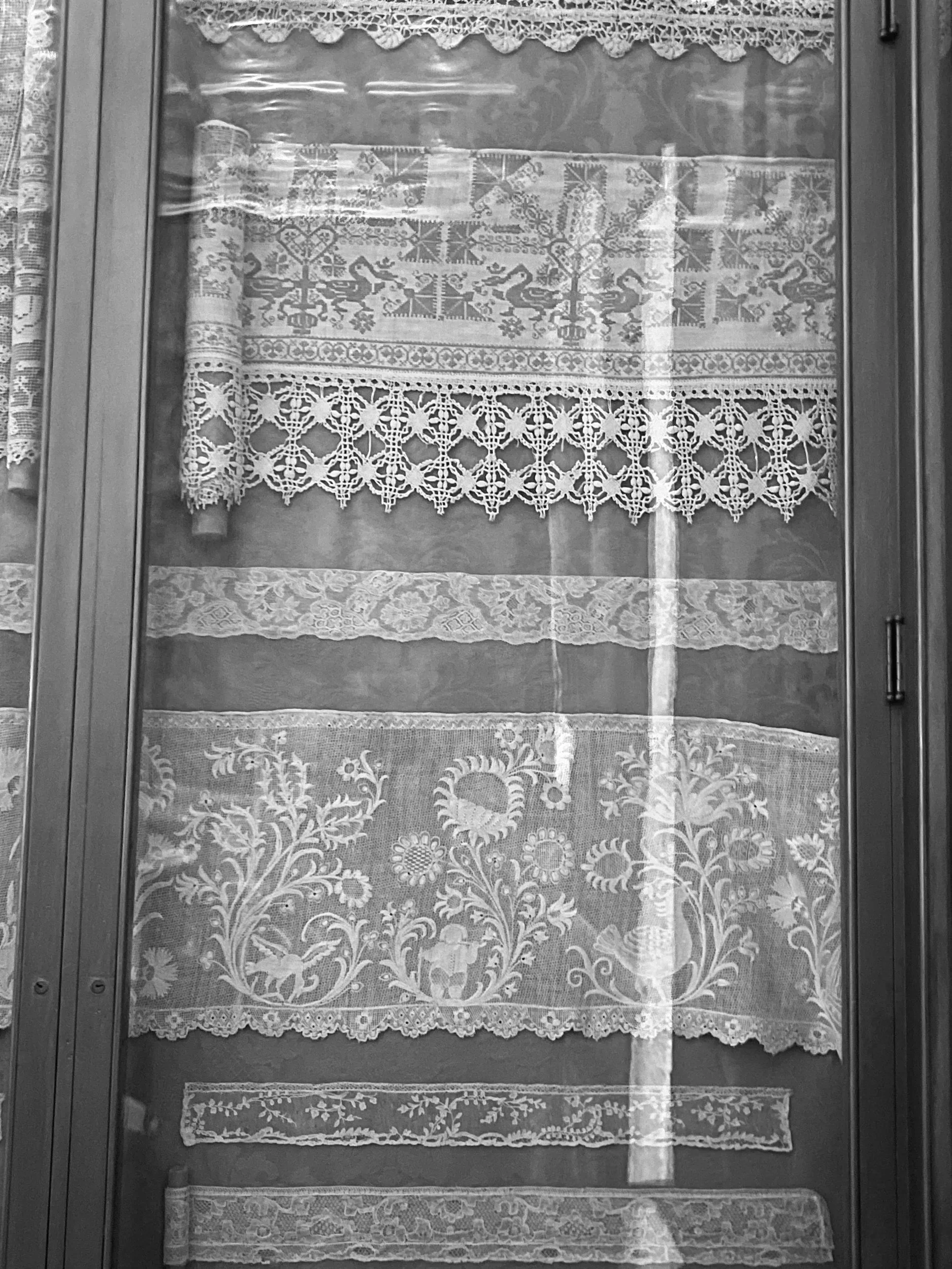 Glass display case containing various pieces of white lace and embroidered fabric. The pieces have intricate patterns, including floral and geometric designs, arranged on different levels inside the case.