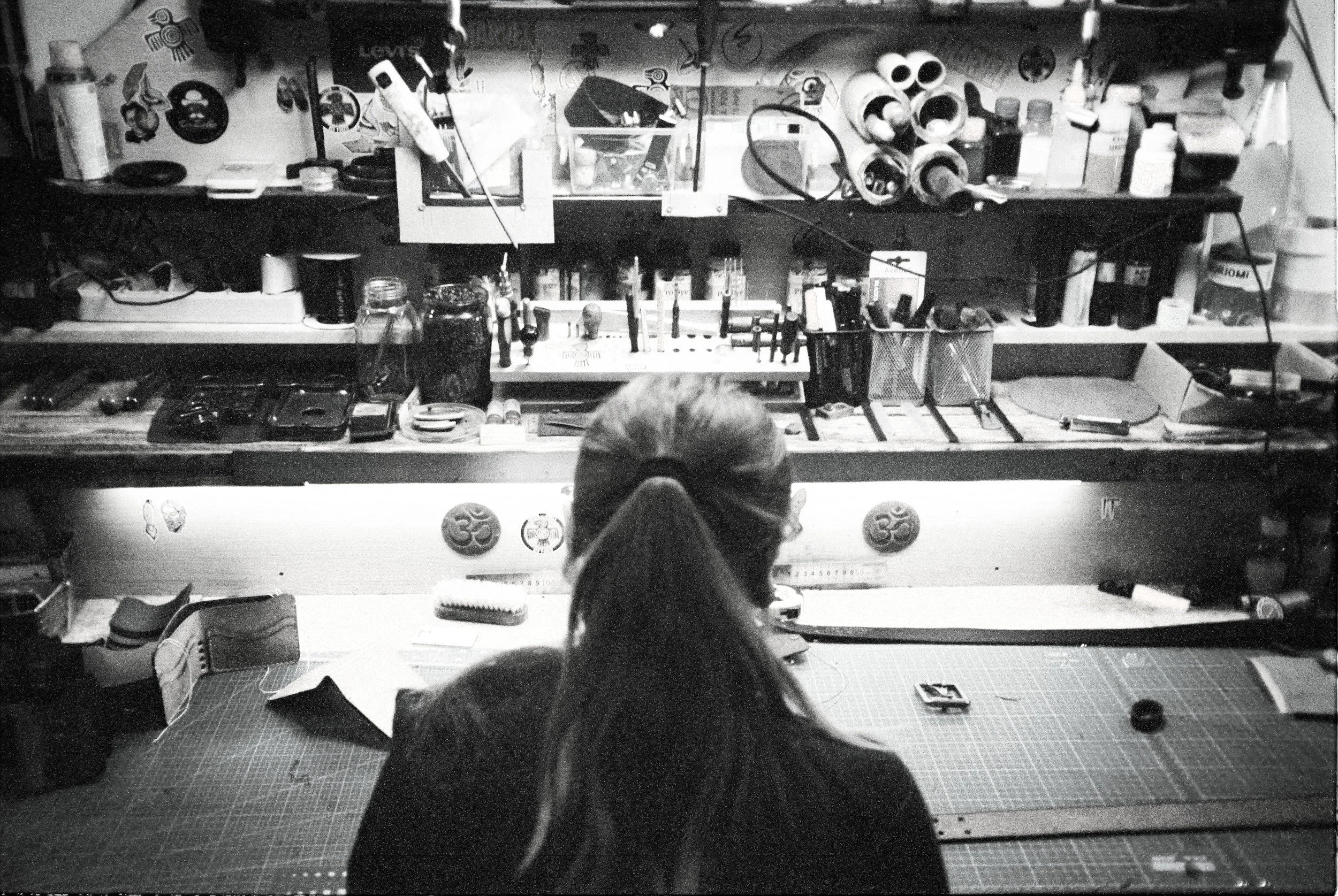 Black and white photo of a cluttered workbench with various tools, bottles, and supplies, viewed from behind a person with short hair wearing glasses, focused on the work area.