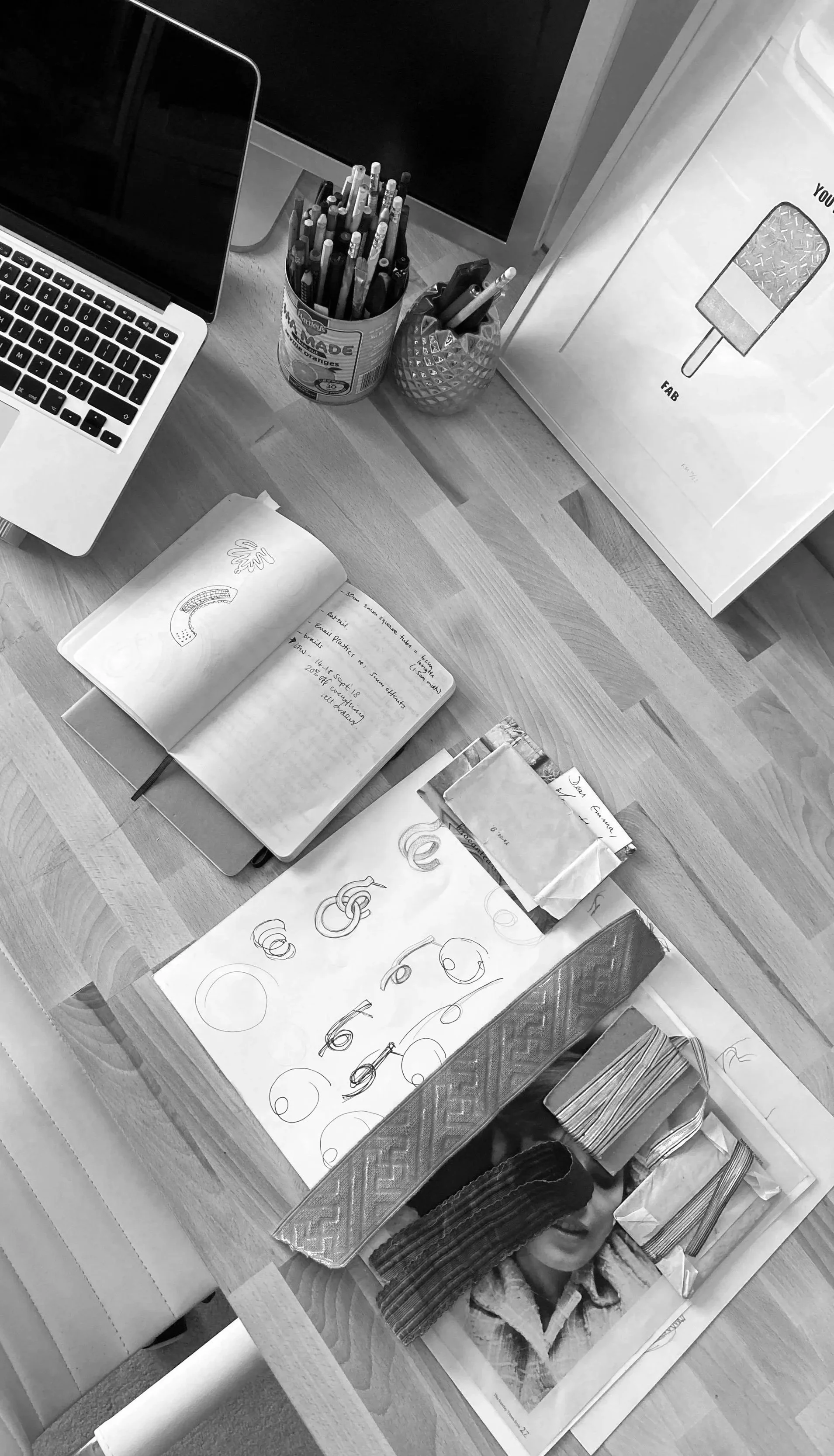 Desk with an open laptop, containers of pens and pencils, framed artwork of a popsicle, sketchbook with handwritten notes and sketches of letters, papers, and photo of a girl with fabric or textured material draped over her face.