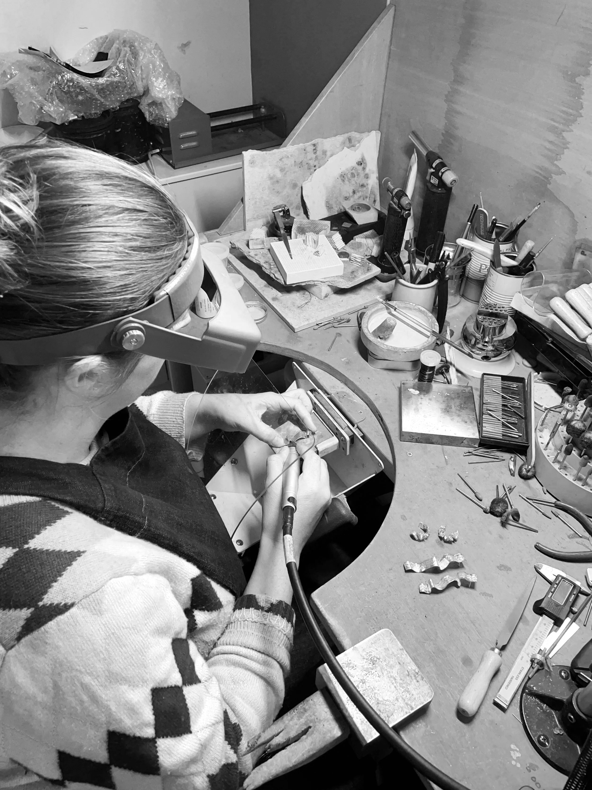 A person wearing glasses and a sweater with a diamond pattern works with small tools at a cluttered workbench, crafting jewelry or small metal items, surrounded by various tools and materials.
