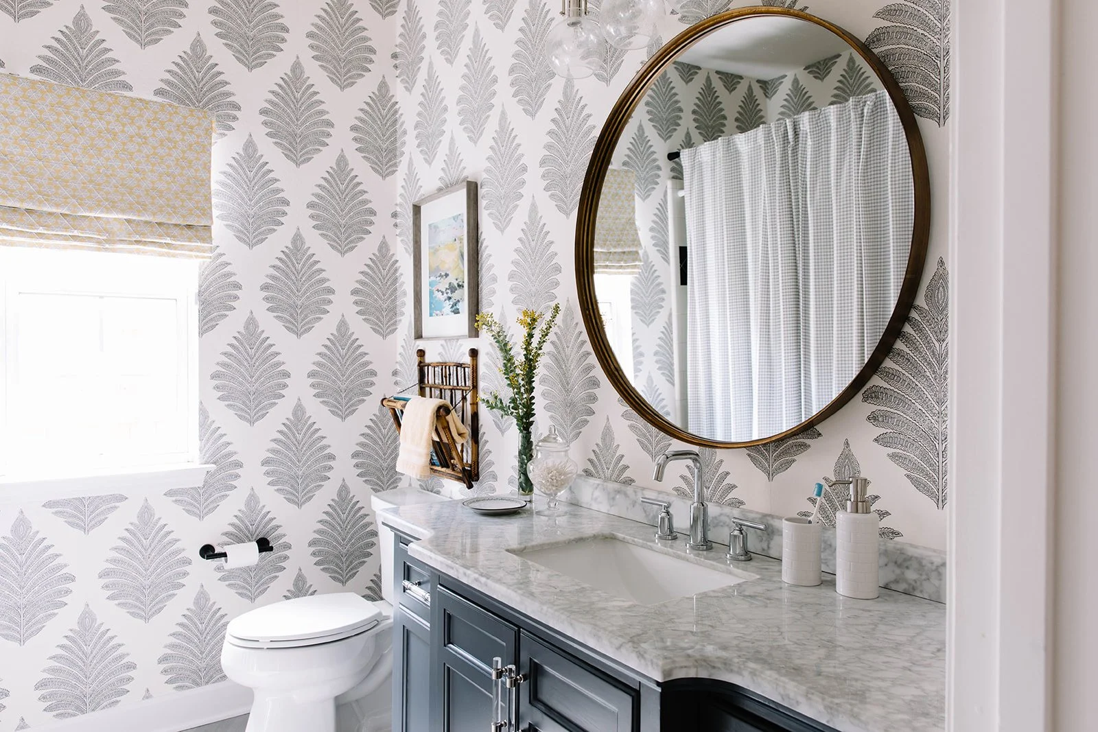 Photo of bathroom with patterned wallpaper. Yellow fabric roman shade on window. Gray vanity with marble countertop. Round mirror with gold frame.