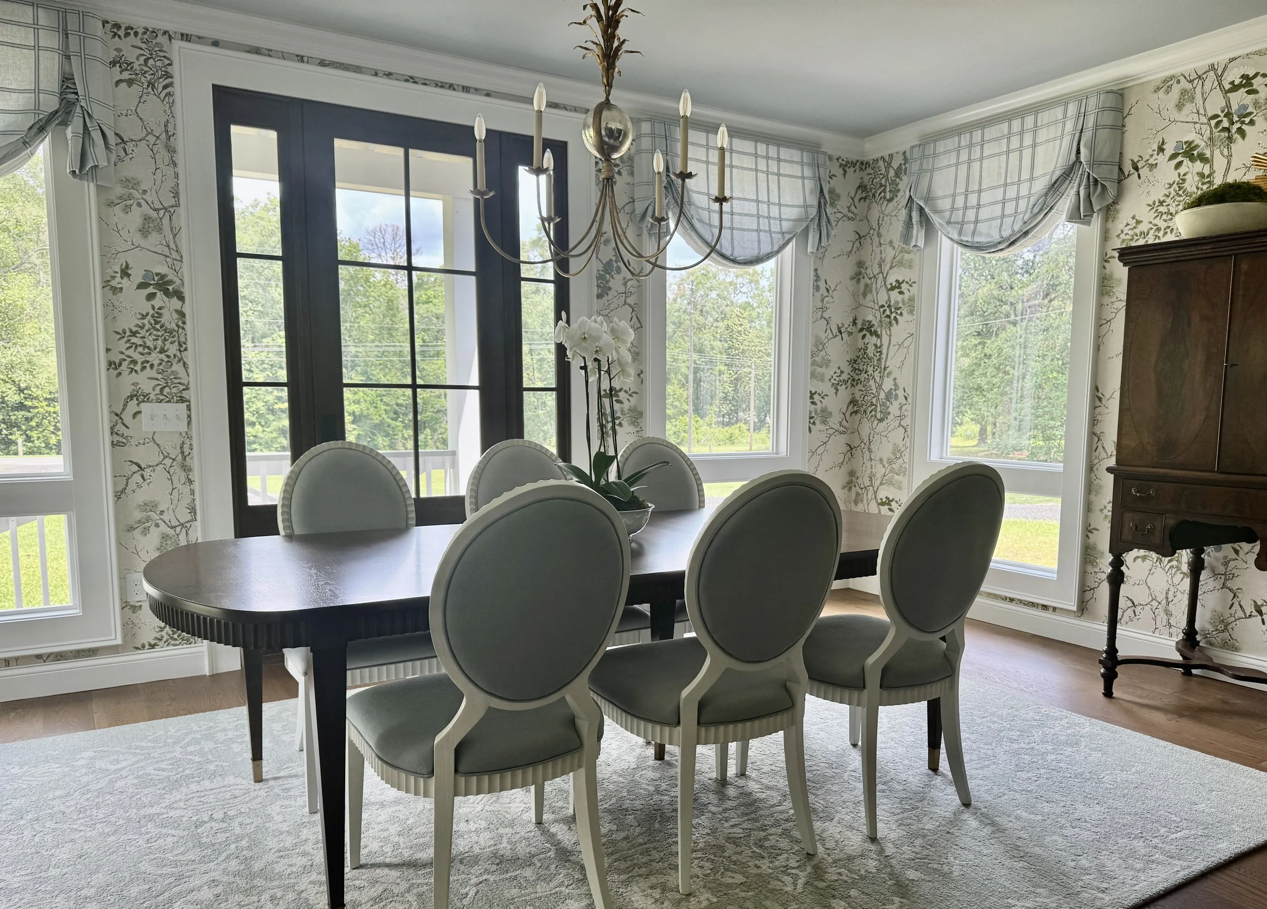 Photo of dining room with patterned wallpaper. Blue valances at the windows. Oval dining table with six white framed chairs. Gold chandelier. Antique tall cabinet.