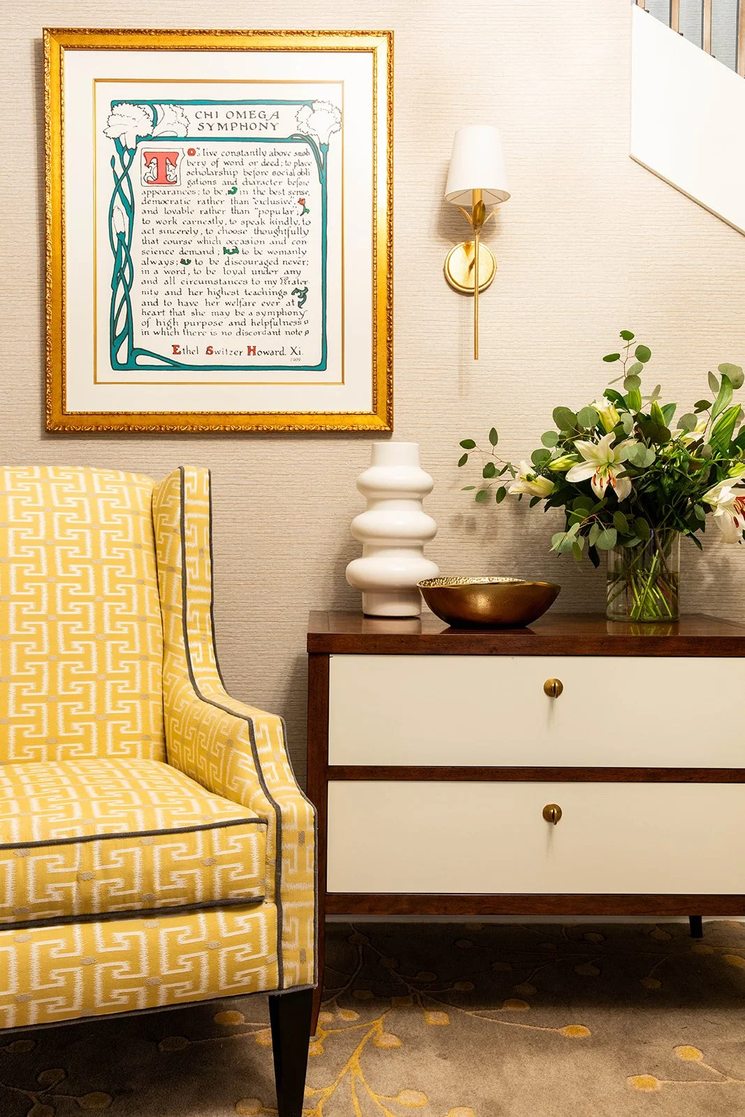 A yellow upholstered chair next to a two-drawer chest with a gold framed print above. A plant and other accessories are on top of the chest. A wall light is mounted above.