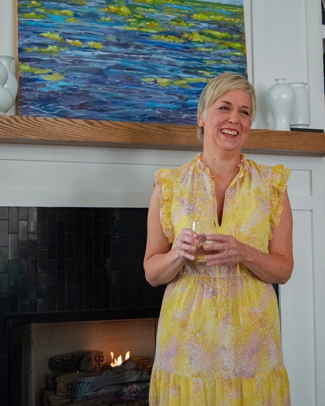 a woman standing in front of a fireplace wearing a yellow dress holding a glass