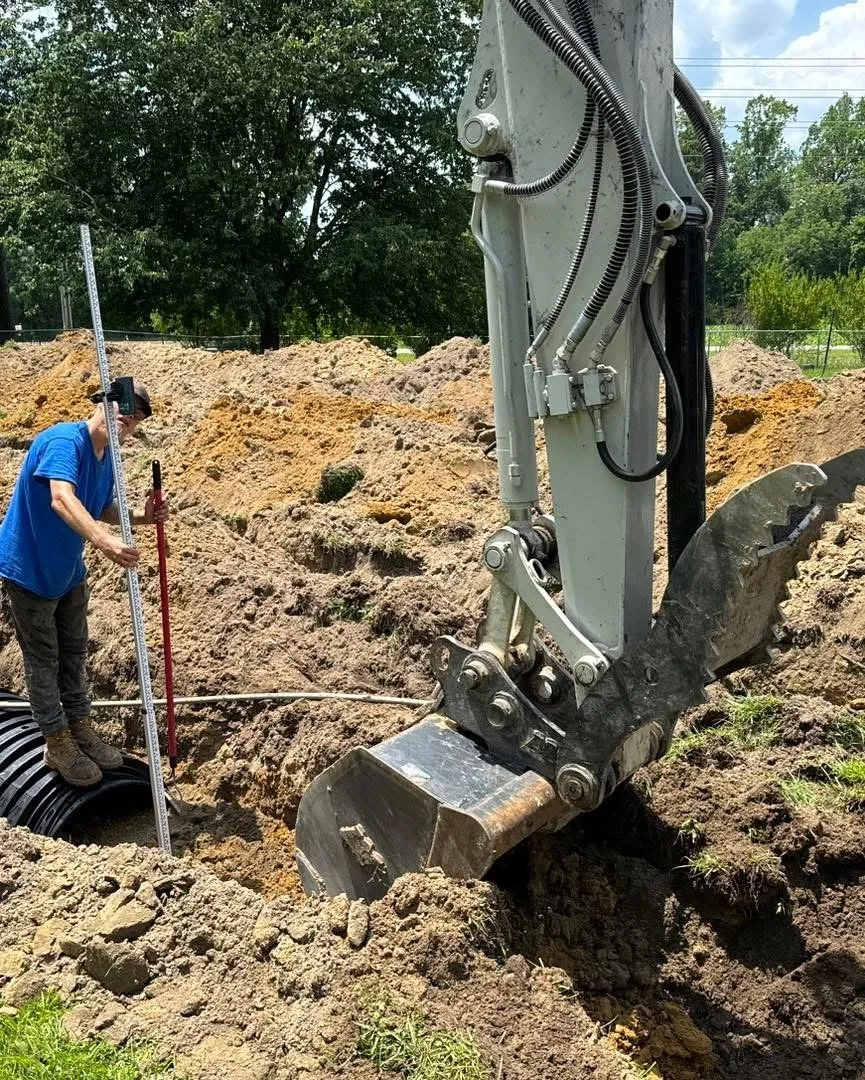 A person in a blue shirt working at a construction site with an excavator digging a trench, surrounded by mounds of dirt and trees in the background.