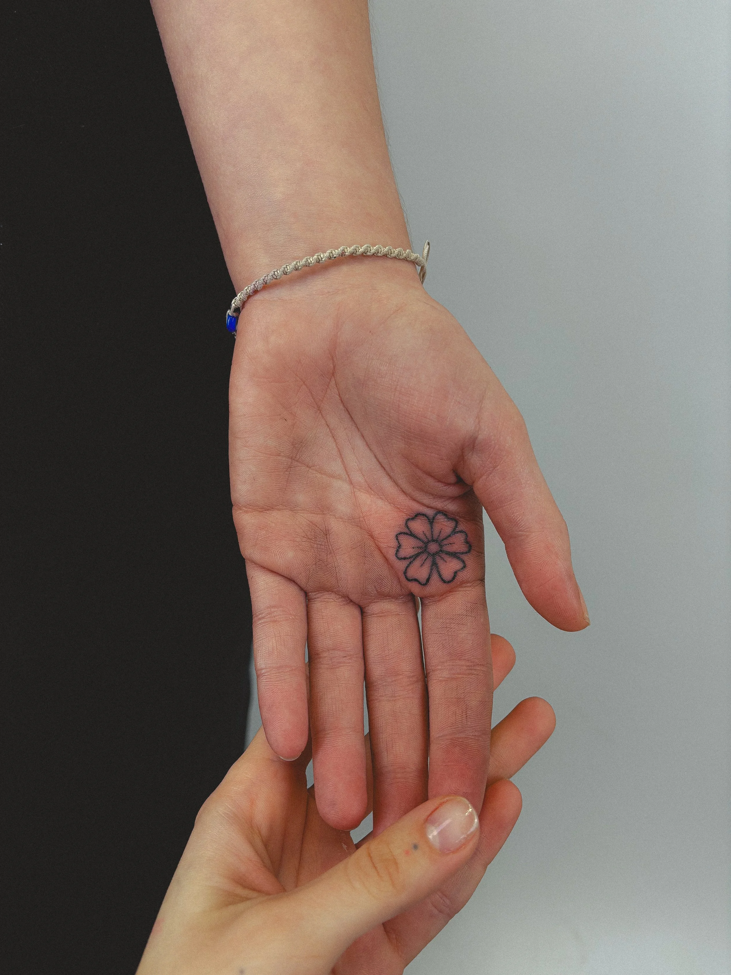 Close-up of two hands, one with a flower tattoo on the wrist, and both wearing a silver bracelet. The background is split into black and gray.