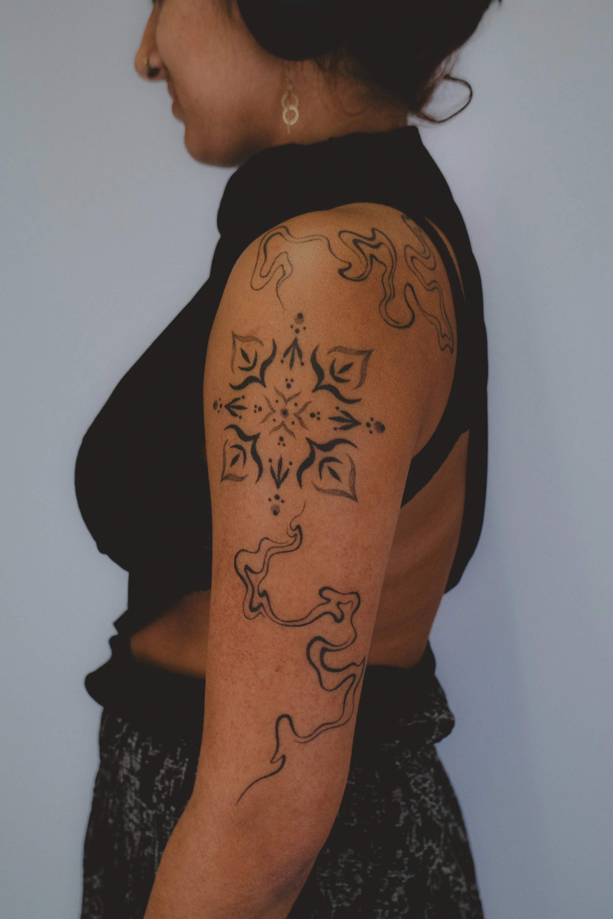 Side view of a woman with tattoos on her arm and shoulder, wearing a black sleeveless top and patterned pants, against a plain light-colored background.