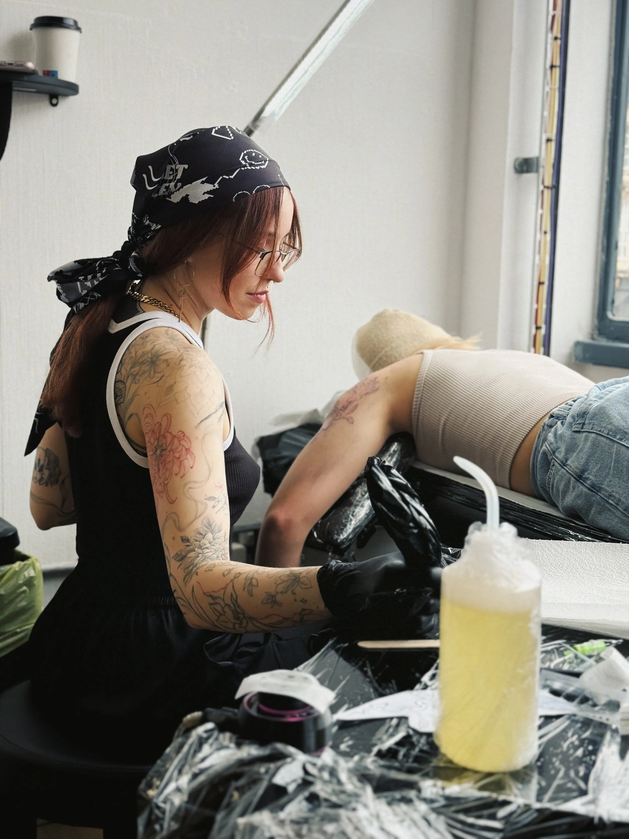 A tattoo artist working on a client's arm in a studio. The artist is wearing glasses, a black bandana, black gloves, and a black sleeveless top. The client is lying face down on a tattoo table, wearing a beige tank top and denim shorts, with tattoos visible on their back and arm.