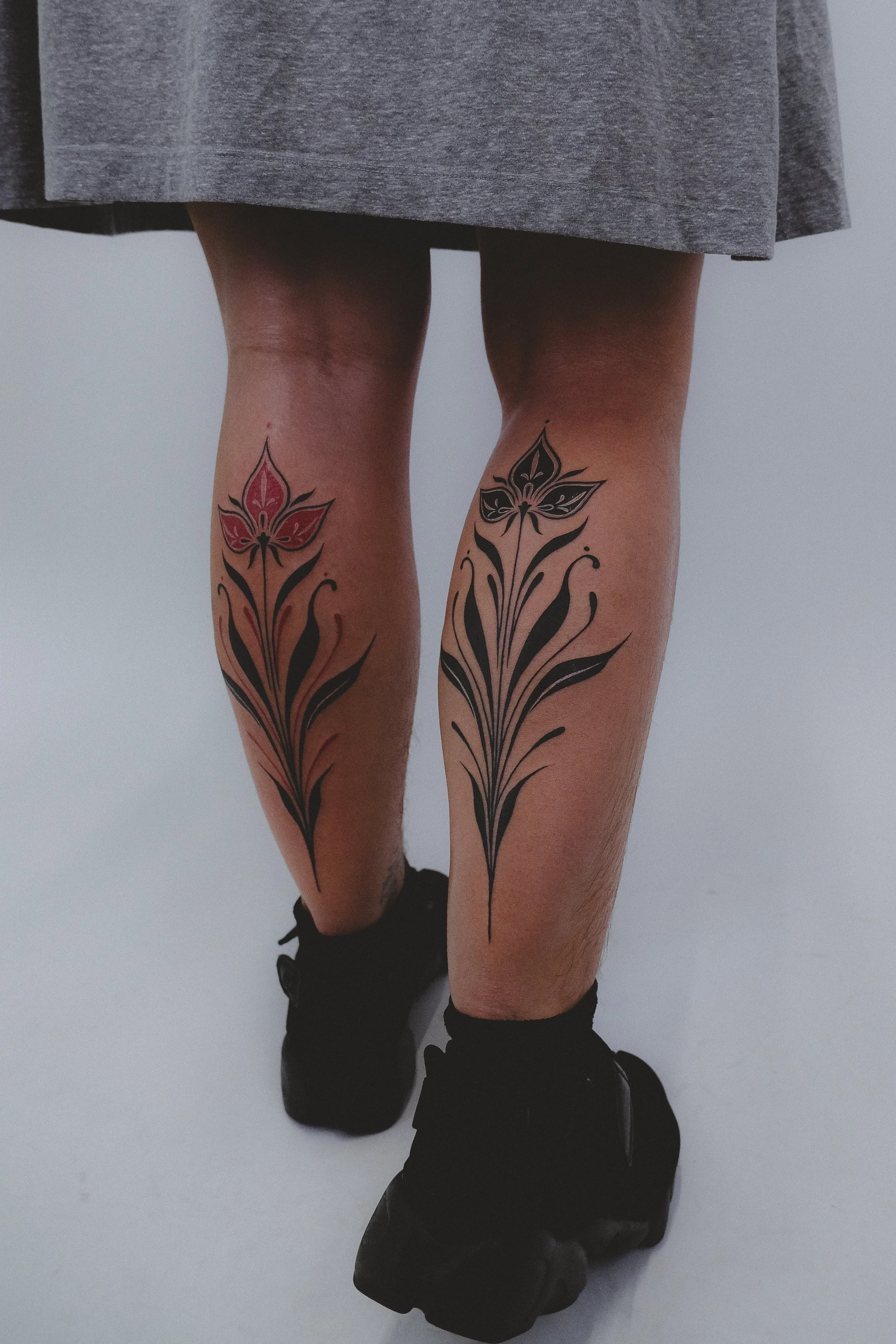 Person wearing black sneakers and black socks, with tattoos of stylized pink and black floral designs on both thighs, standing on a light-colored floor with a plain background.
