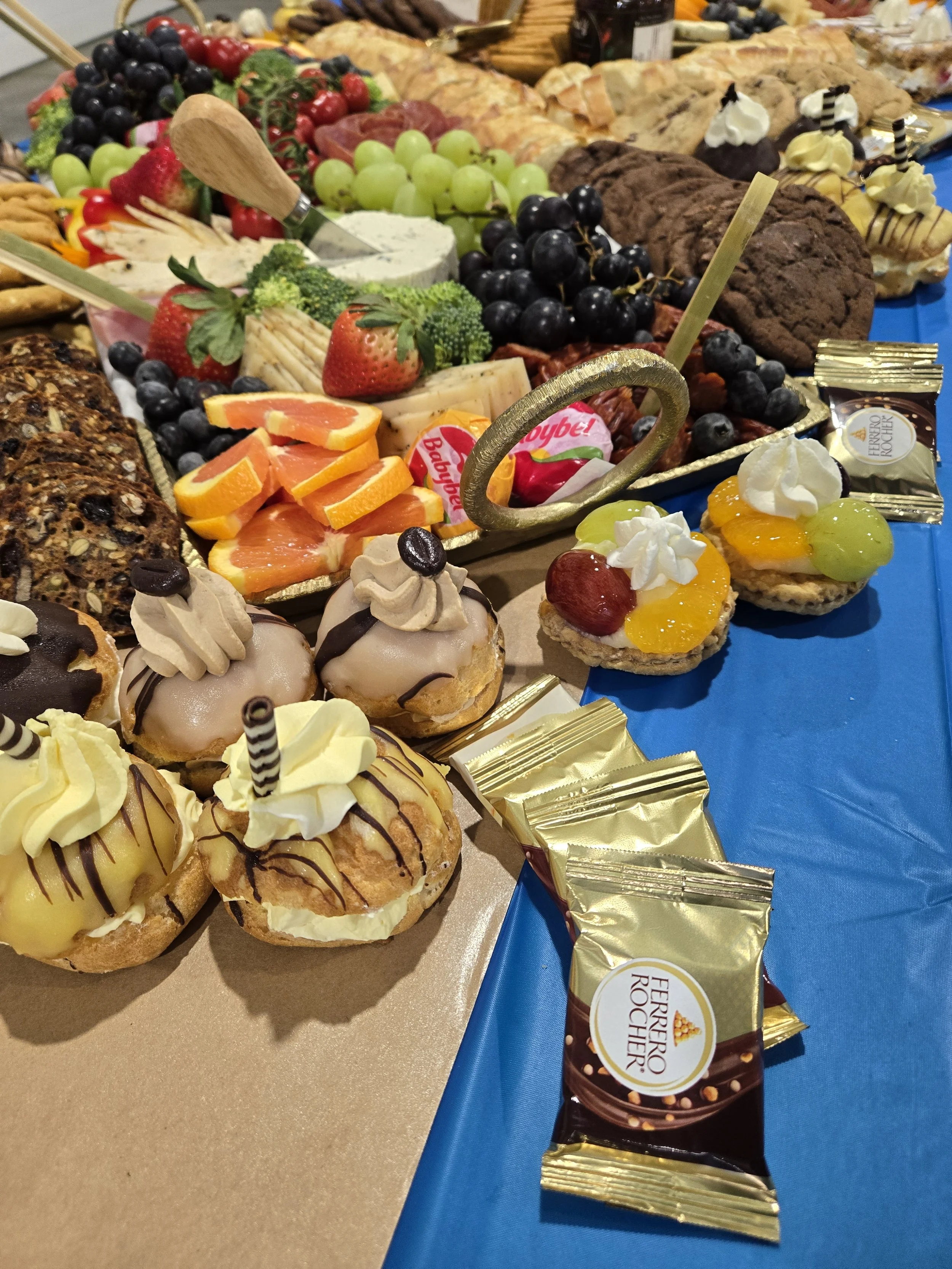 A dessert table with various pastries, chocolates, and fruit. Close-up of profiteroles topped with whipped cream and chocolate, alongside small fruit-topped tarts with grapes, cherries, and peaches. In the background, there are cookies, candies, grap