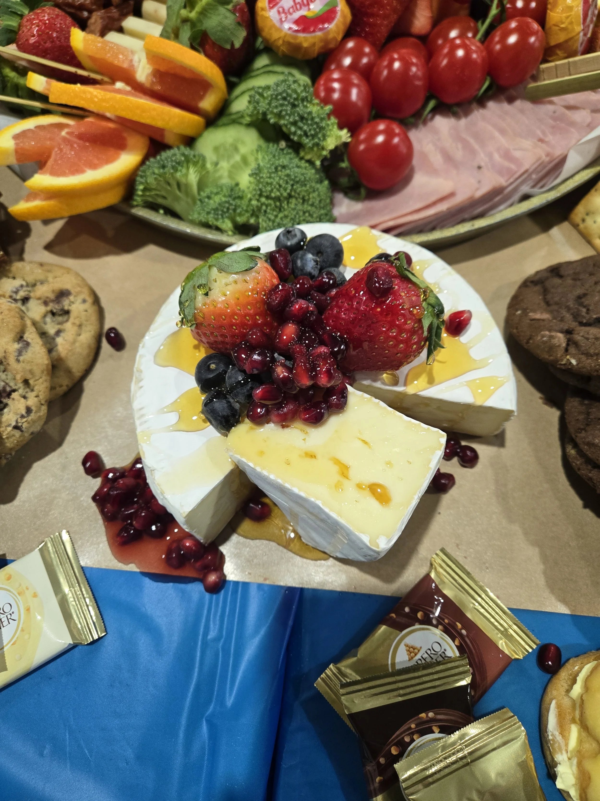 A variety of fresh fruits including strawberries, blueberries, blackberries, pomegranate seeds, and sliced citrus on a serving plate with a wheel of brie cheese topped with berries and honey, surrounded by cookies, chocolates, and a platter of vegeta
