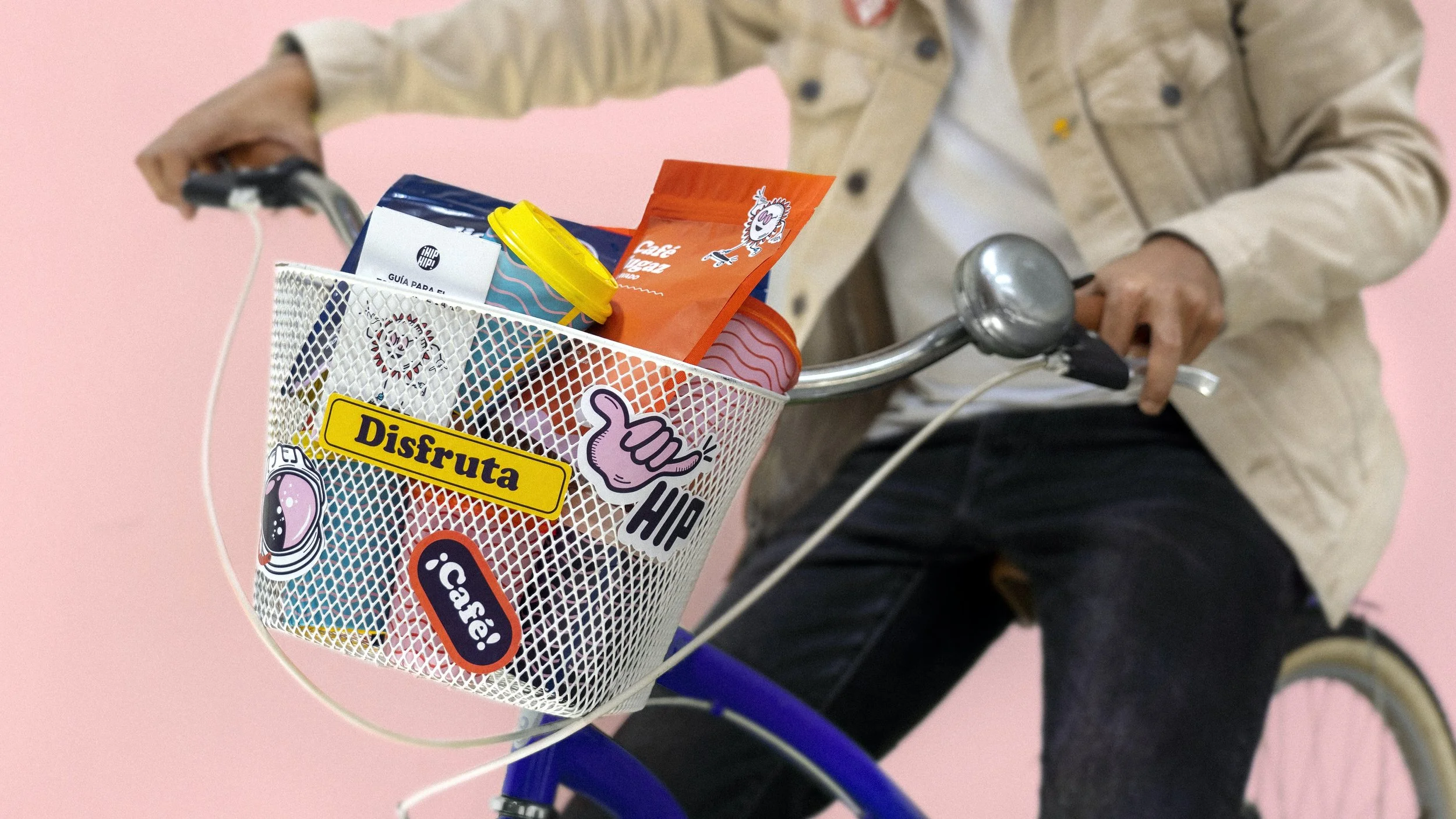 A person riding a bicycle with a white basket on the front, filled with snacks and beverages, against a pink background.