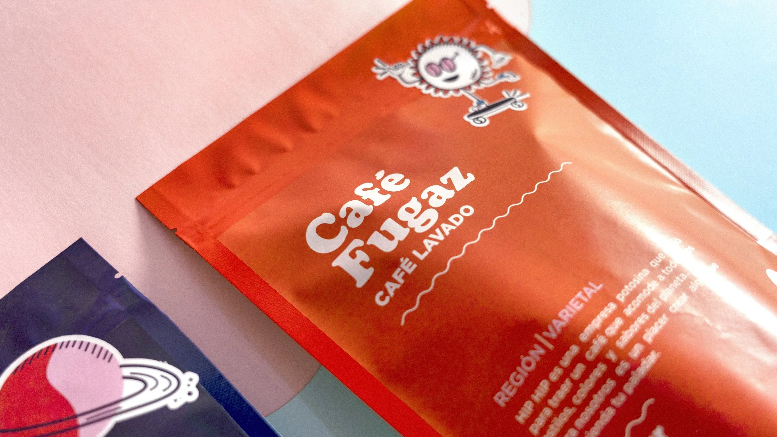 Close-up of a red package with the text "Café Fulgaz" and "Café Lavado," featuring a cartoon character skateboarding at the top. Part of a blue package with a graphic and a pink surface in the background.