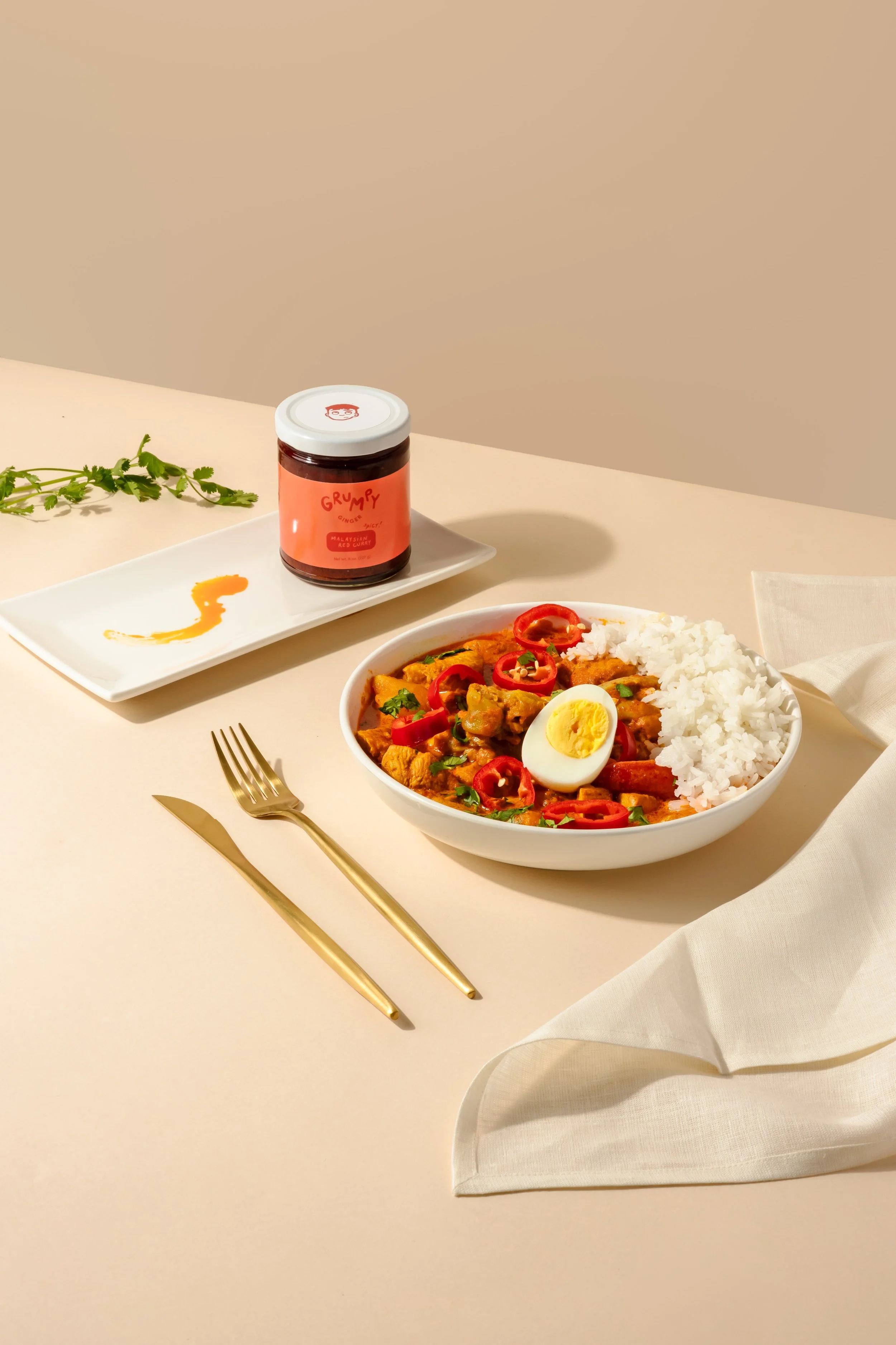 A bowl of rice with curry, garnished with sliced red chili peppers, a boiled egg, and green herbs, on a light-colored table. Beside it are gold-colored fork and knife, with a white napkin, a small white rectangular plate with a smear of curry sauce, a small jar of curry sauce labeled "Grumpy" with a white lid, and some green herbs.