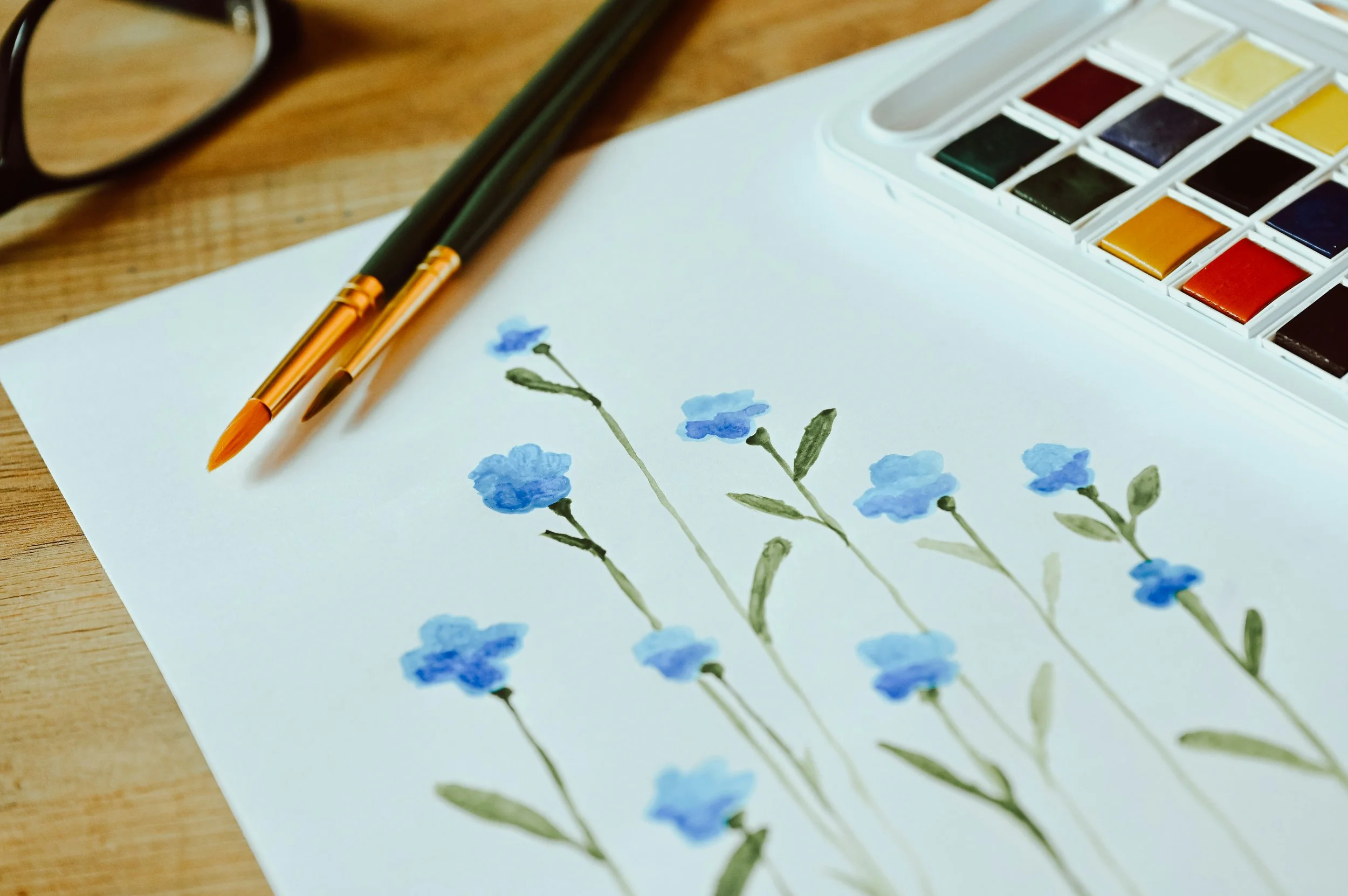 Watercolour paints and brushes beside a simple blue floral painting.
