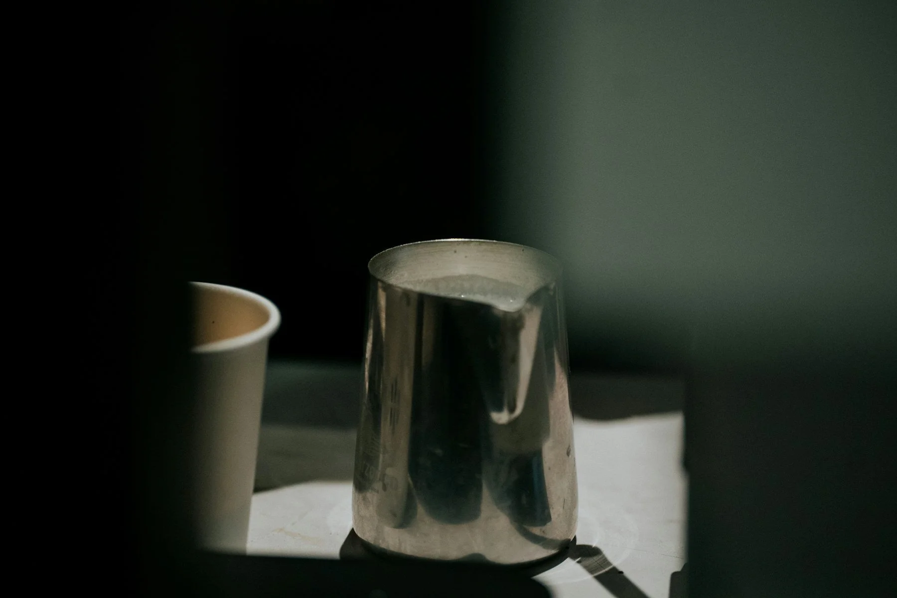 Stainless steel pitcher lit by strong sunlight beside a paper cup