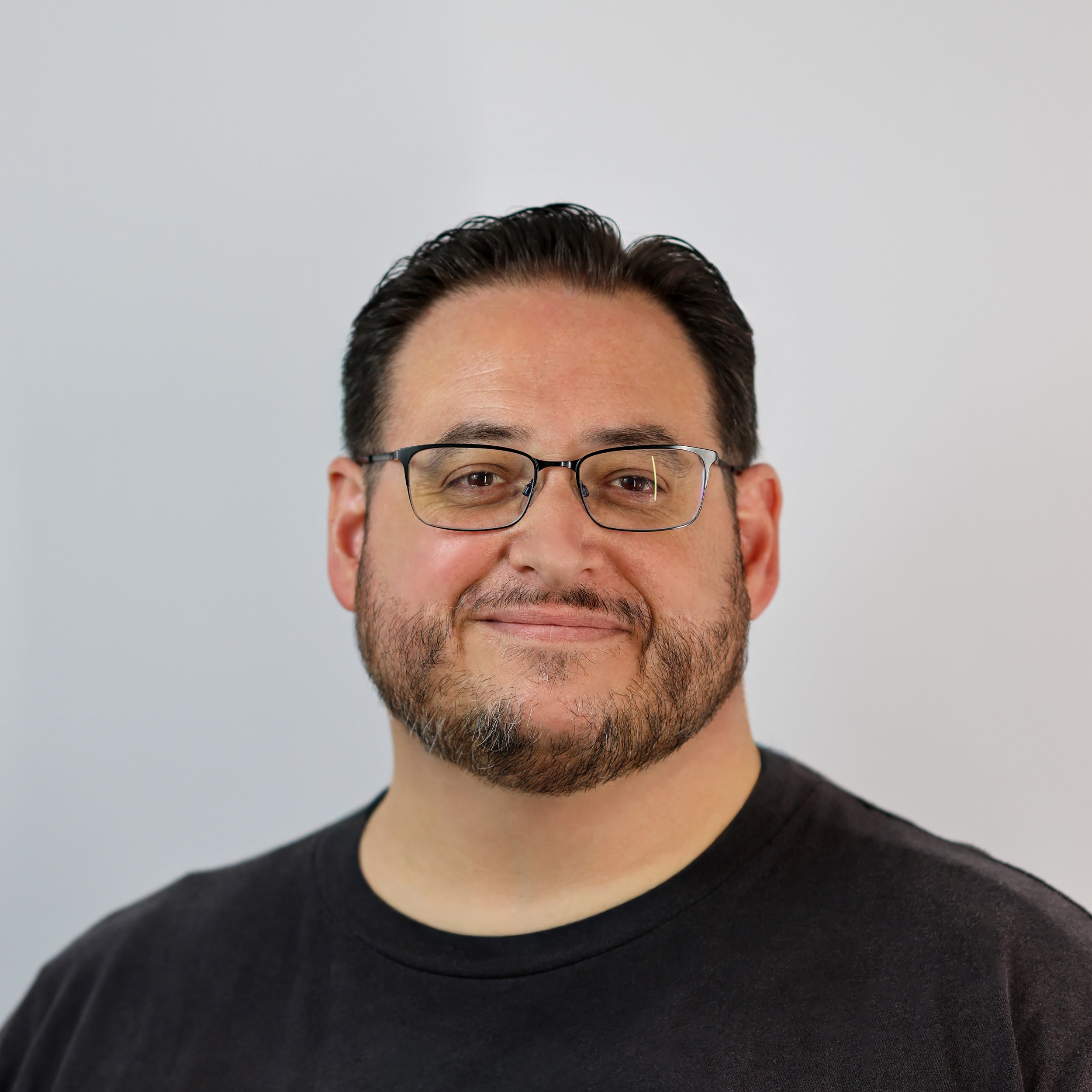 Man with glasses smiling, wearing a black shirt, standing indoors with a blurred painting in the background.