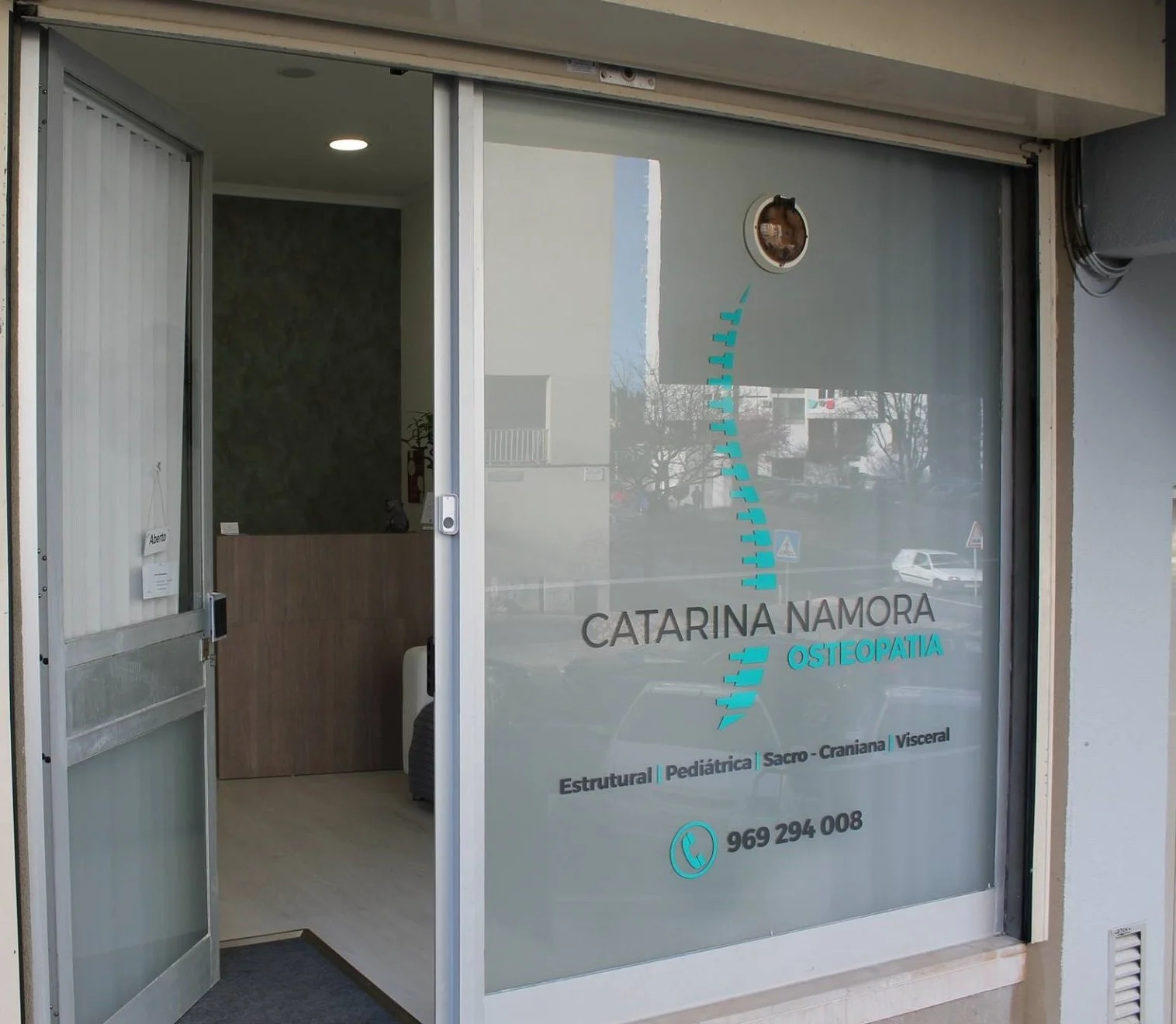 Fachada de uma clínica de osteopatia com vitrine de vidro, nome 'Catarina Namora Osteopatia', que oferece tratamentos estruturais, pediátricos, sacro-cranianos e viscerais, e telefone de contato. A porta está aberta, revelando o interior da clínica.