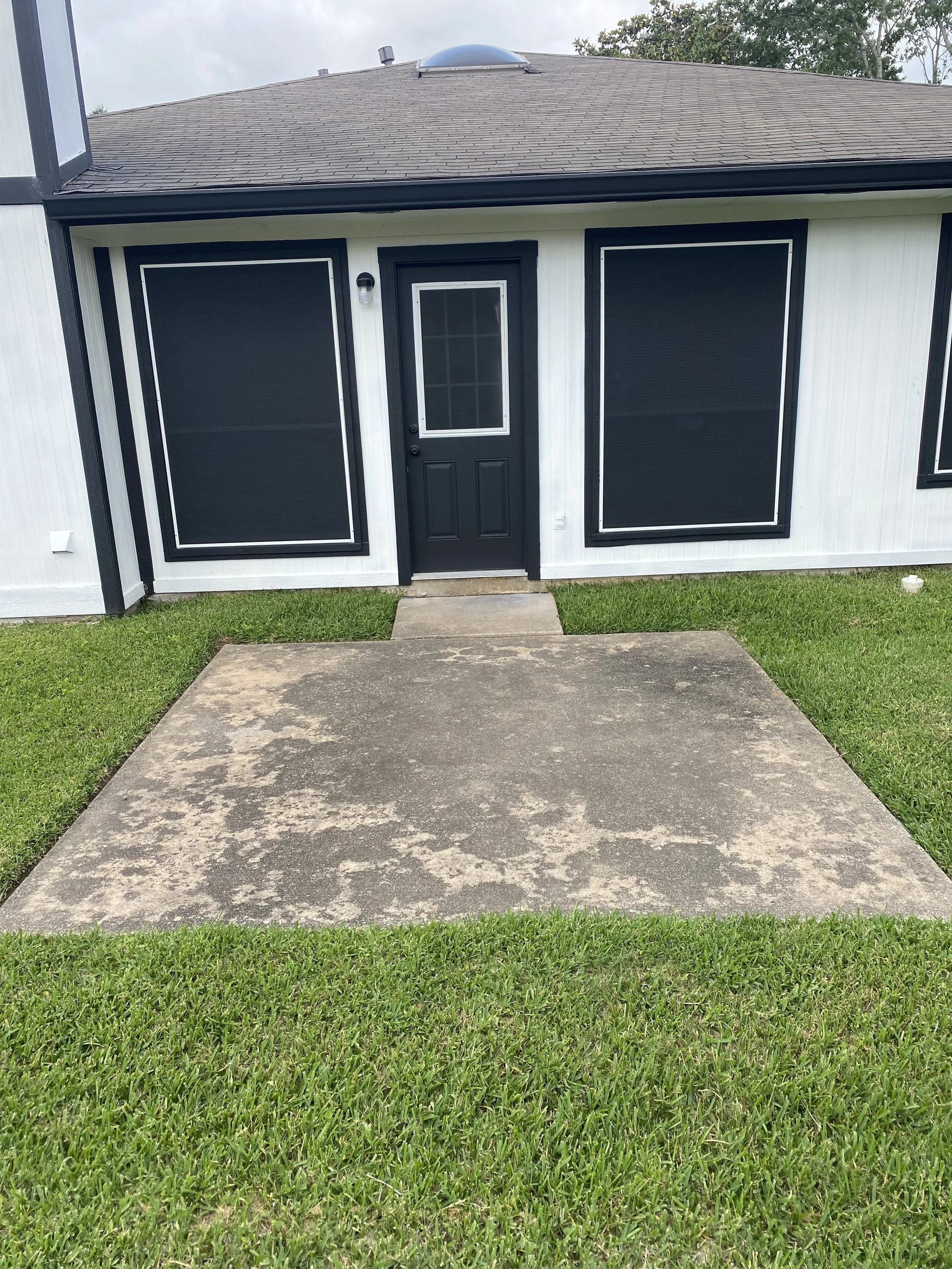 Back patio with stained and weathered concrete before pressure washing, showing dark discoloration and patchy buildup against a white and black-trimmed house in Houston, TX