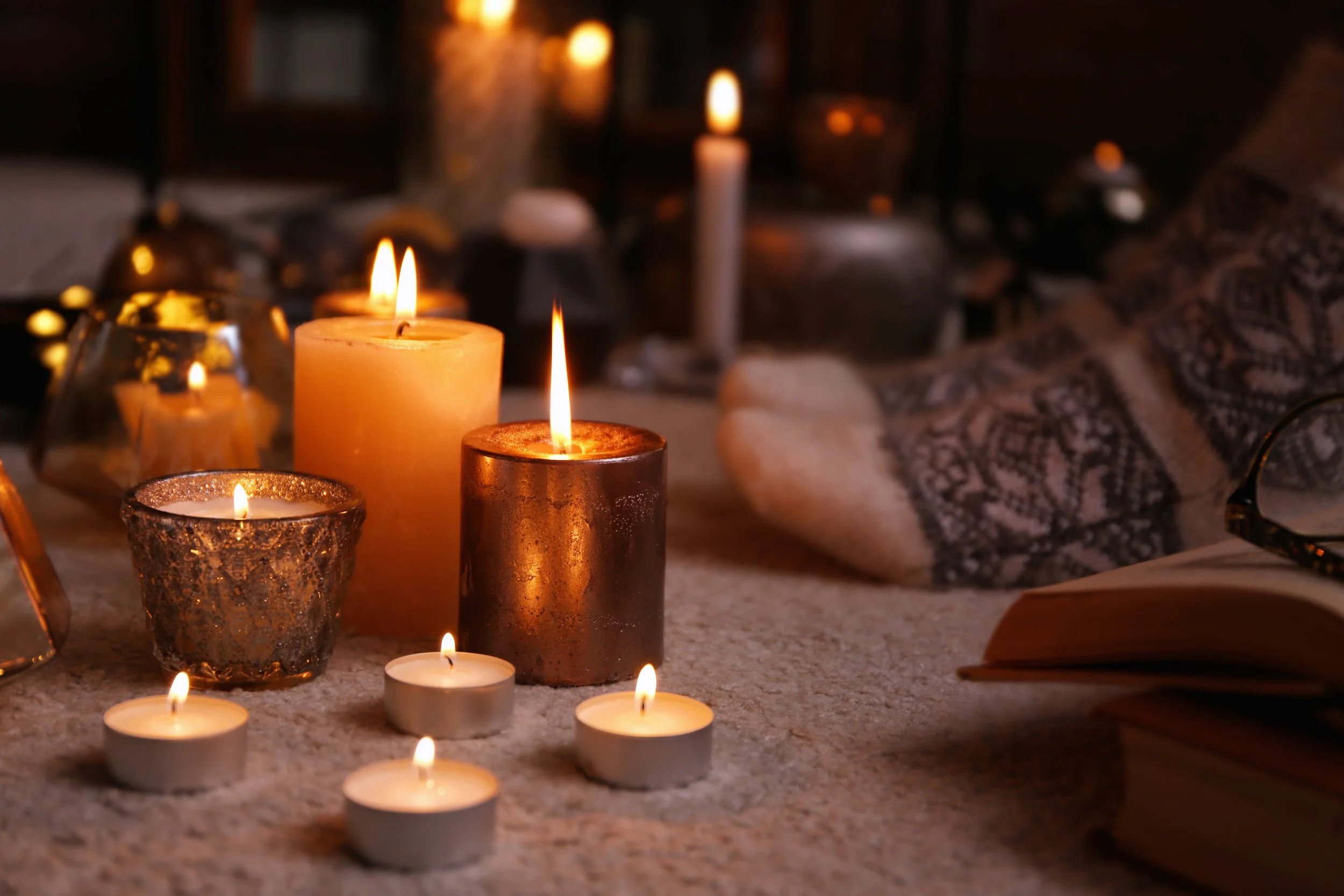 Various lit candles of different sizes and colors placed on a carpeted surface, with an open book, glasses, and a patterned cloth nearby, creating a cozy and warm ambiance.