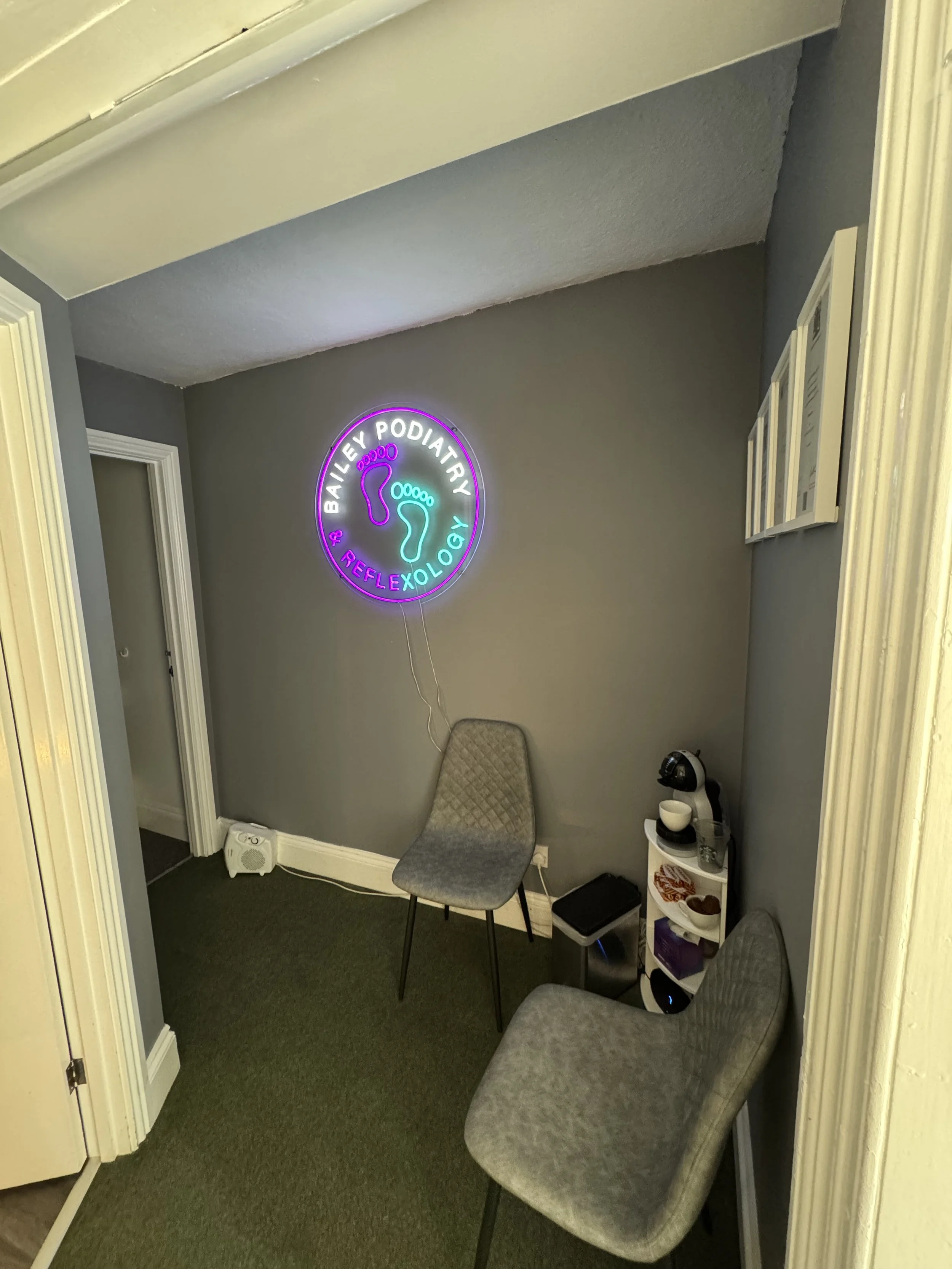 Interior of a room with two gray chairs, a small white shelf with a coffee machine, and a neon sign on the wall that says 'Bailey Podiatry & Reflexology' with footprints in neon lights.