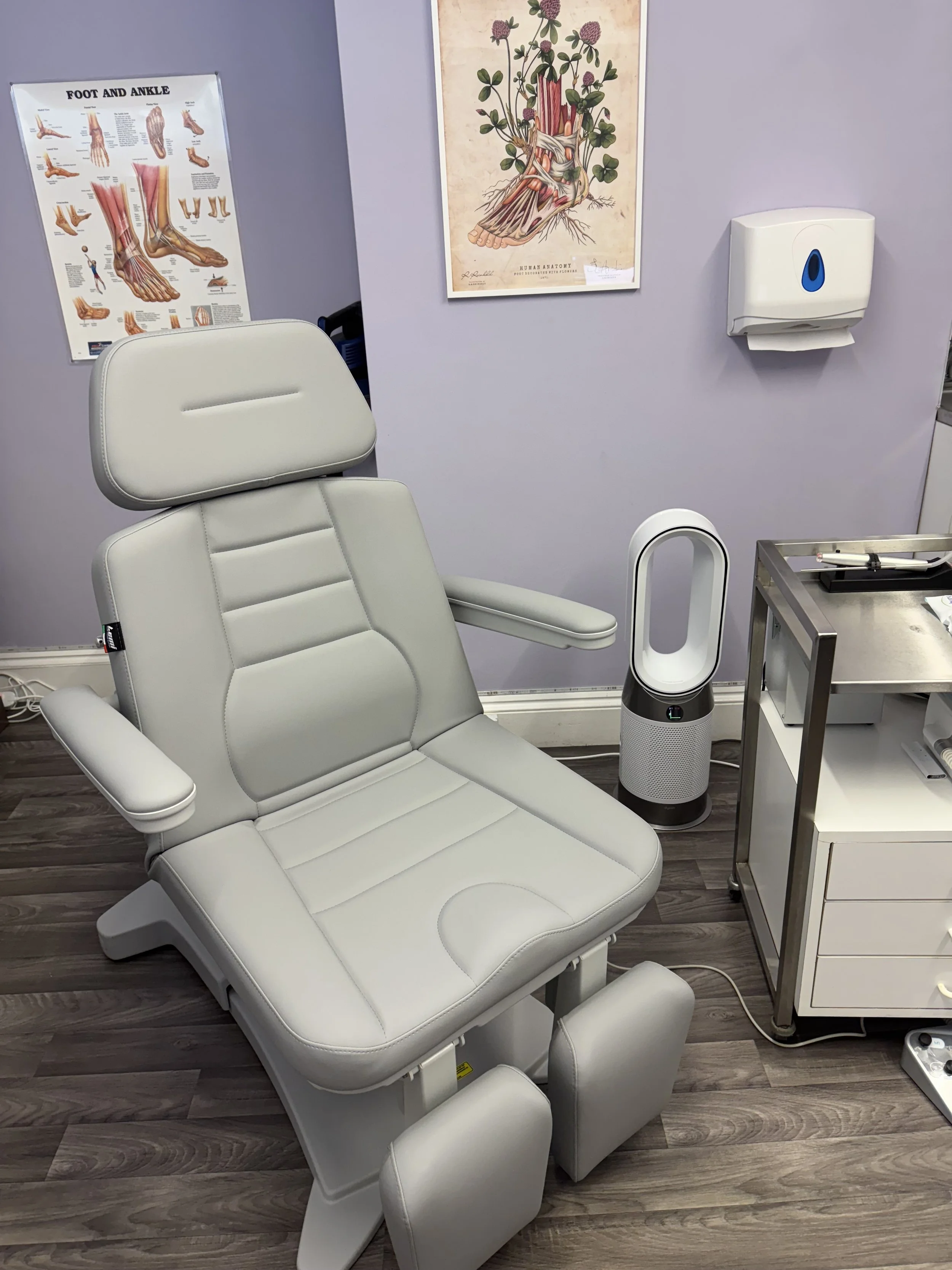 Medical examination chair in a clinic room with anatomical posters, a Dyson air purifier, and medical supplies.