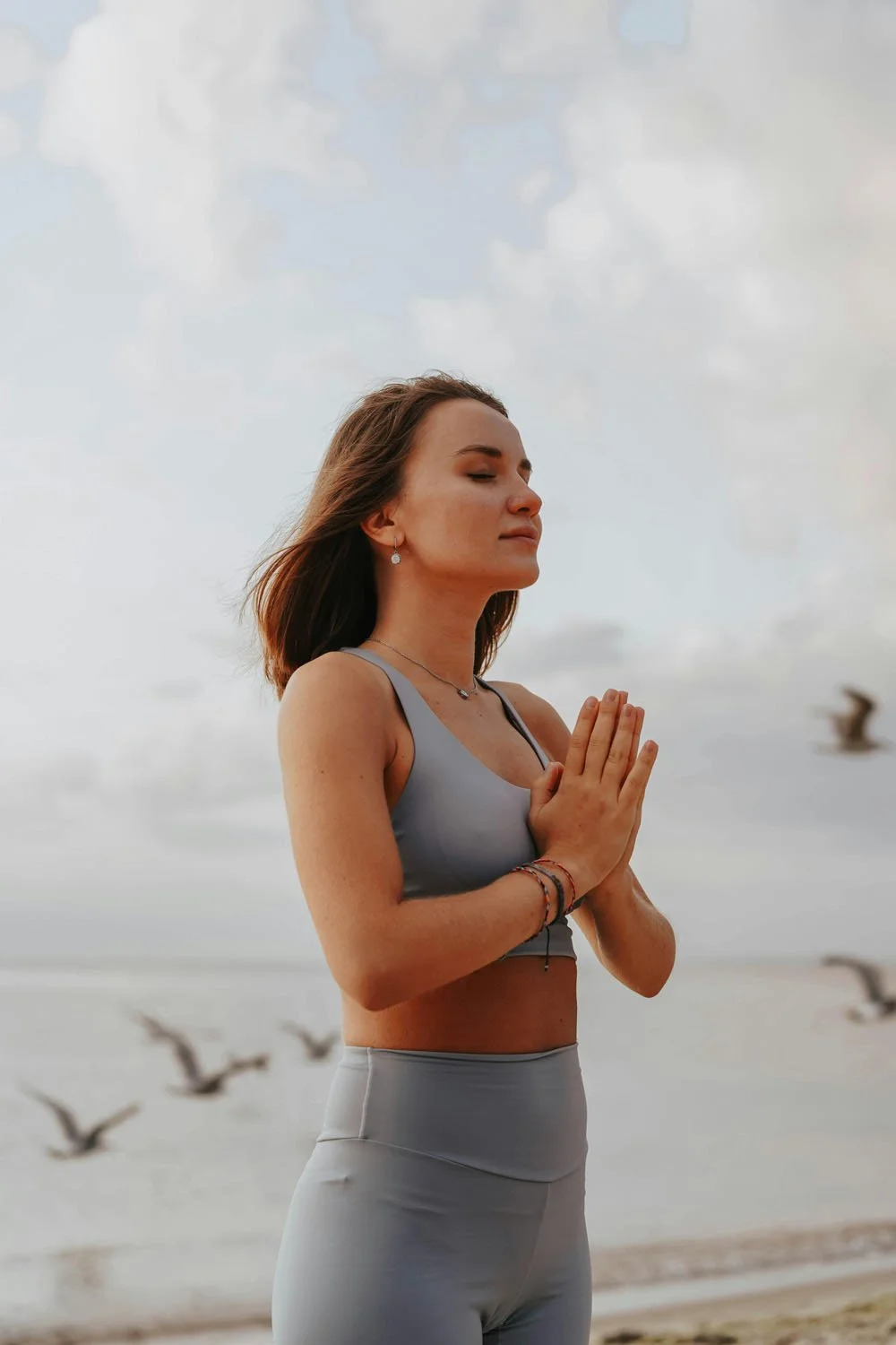 Woman meditating with hands in prayer pose on a windswept beach.