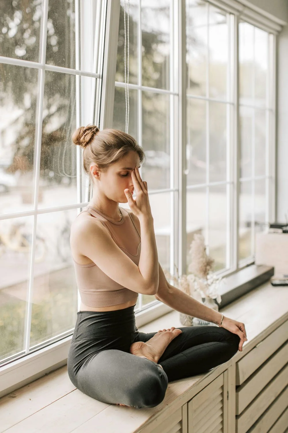 Woman practicing alternate nostril breathing beside a sunlit window.