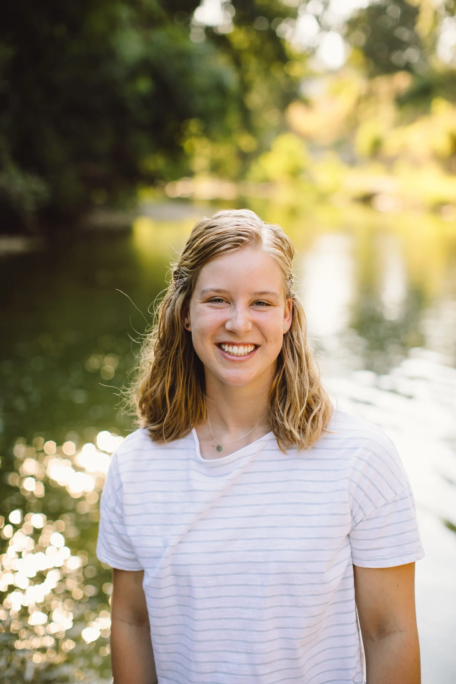 Smiling young woman with wavy blonde hair standing outdoors near a body of water with trees in the background.